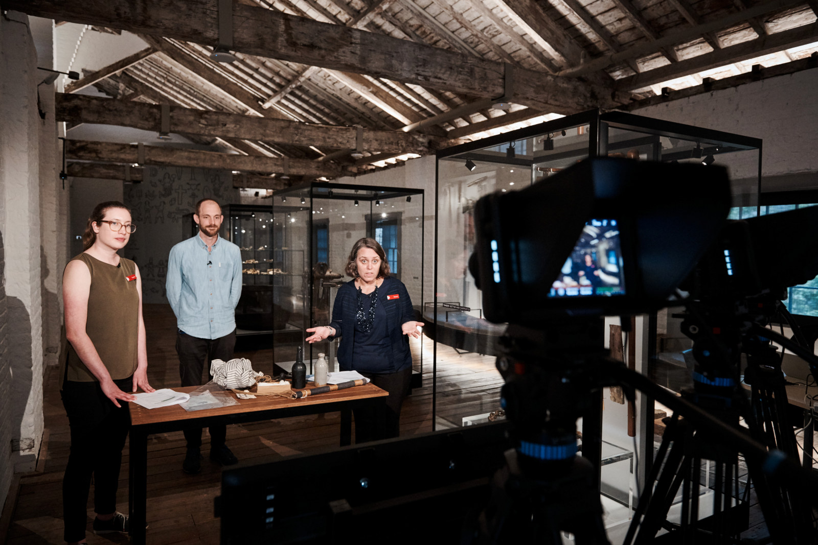 Sophie Reid, Producer – Learning Programs; Carlin de Montfort, curator; and Naomi Manning, Producer – Learning Programs, stand in front of a series of cameras as they present a virtual excursion. They are standing behind a table of historical objects in the Meet the Convicts room on the top floor of the Hyde Park Barracks.
