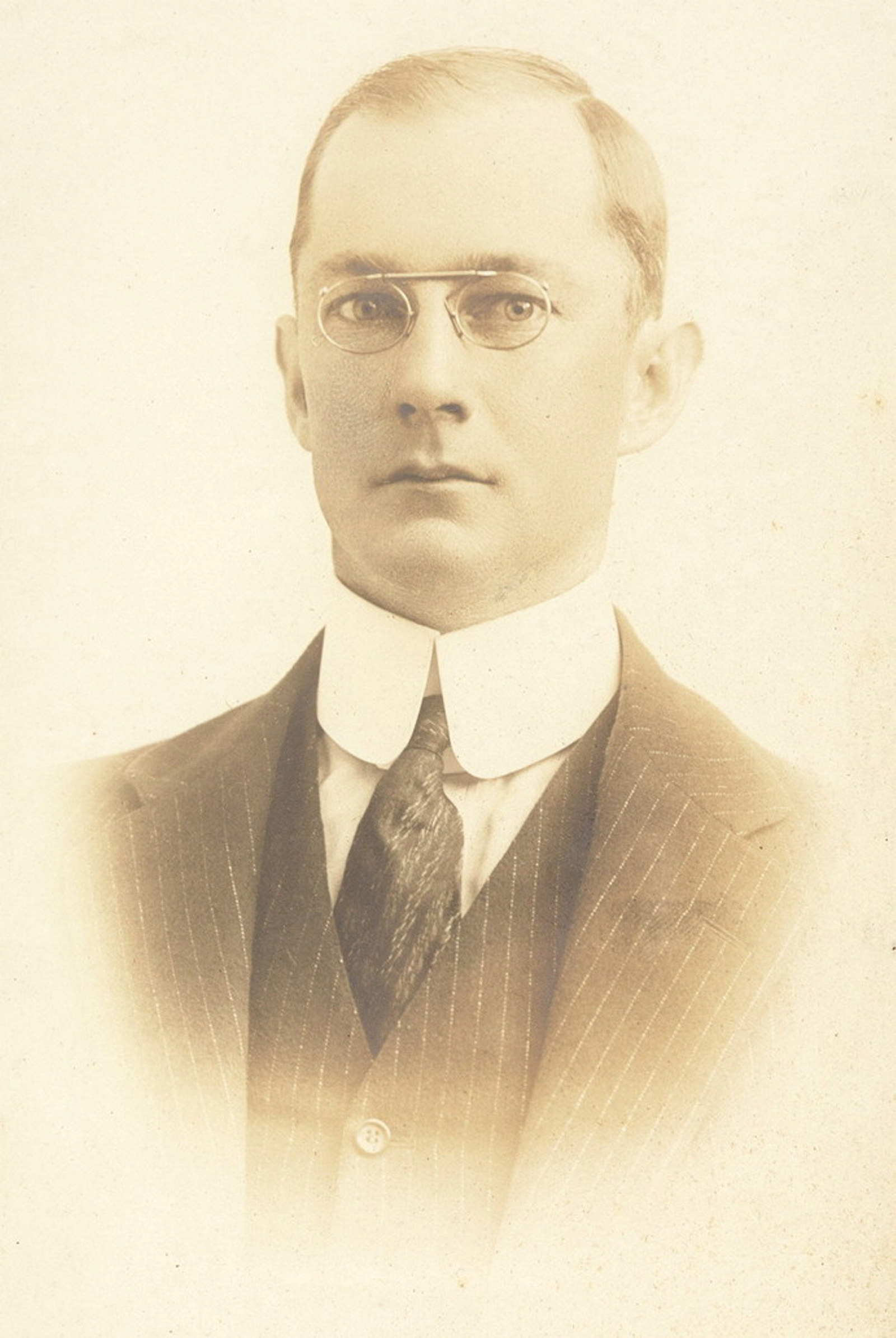 Sepia toned photo of man wearing glasses with a coat, high collar and tie.