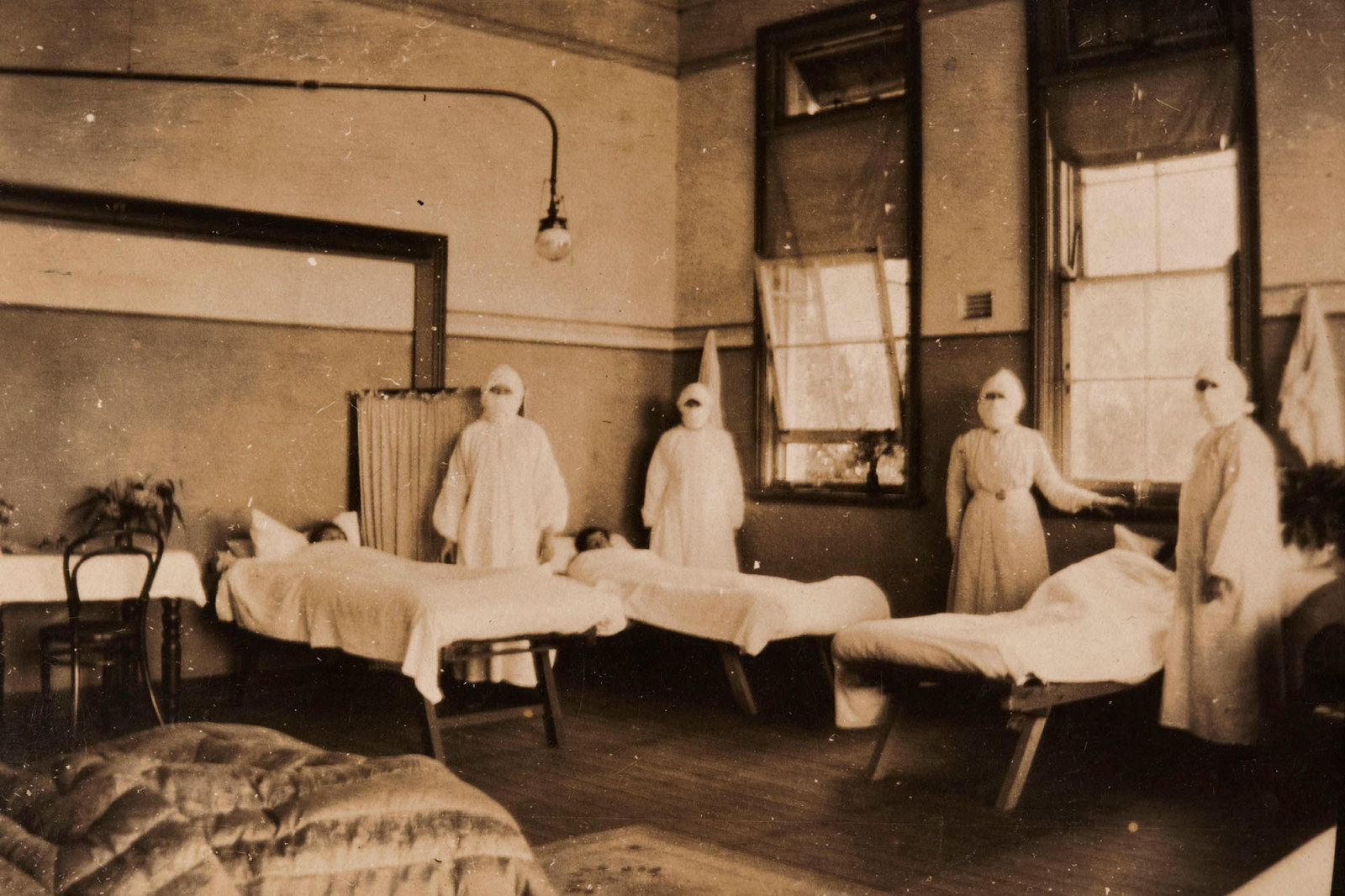 Four women in masks and nurse uniforms in room set up with hospital beds.