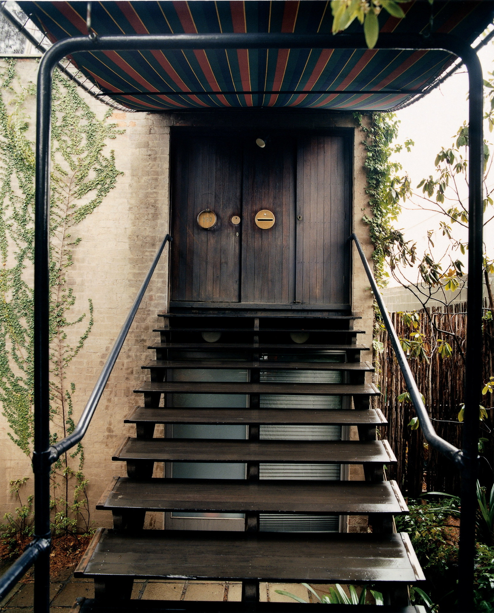This is a colour photograph of open shallow timber staircase leading to a large timber door with a canopy covering the stairs and doorway