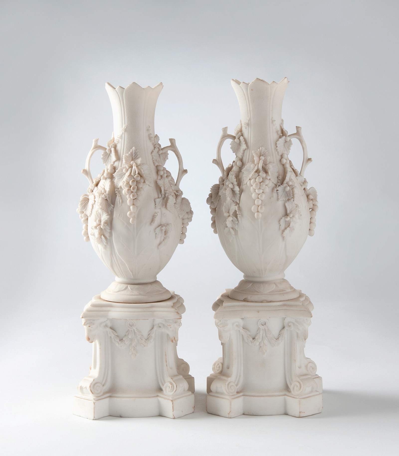 Shaped pedestal base with corners decorated with scrolls to the foot and ram's heads to the upper edge liked by applied swags; the vase with circular flaring foot, ovoid body and long, narrow neck, rim with pointed edge. Pair of handles in the form of bra