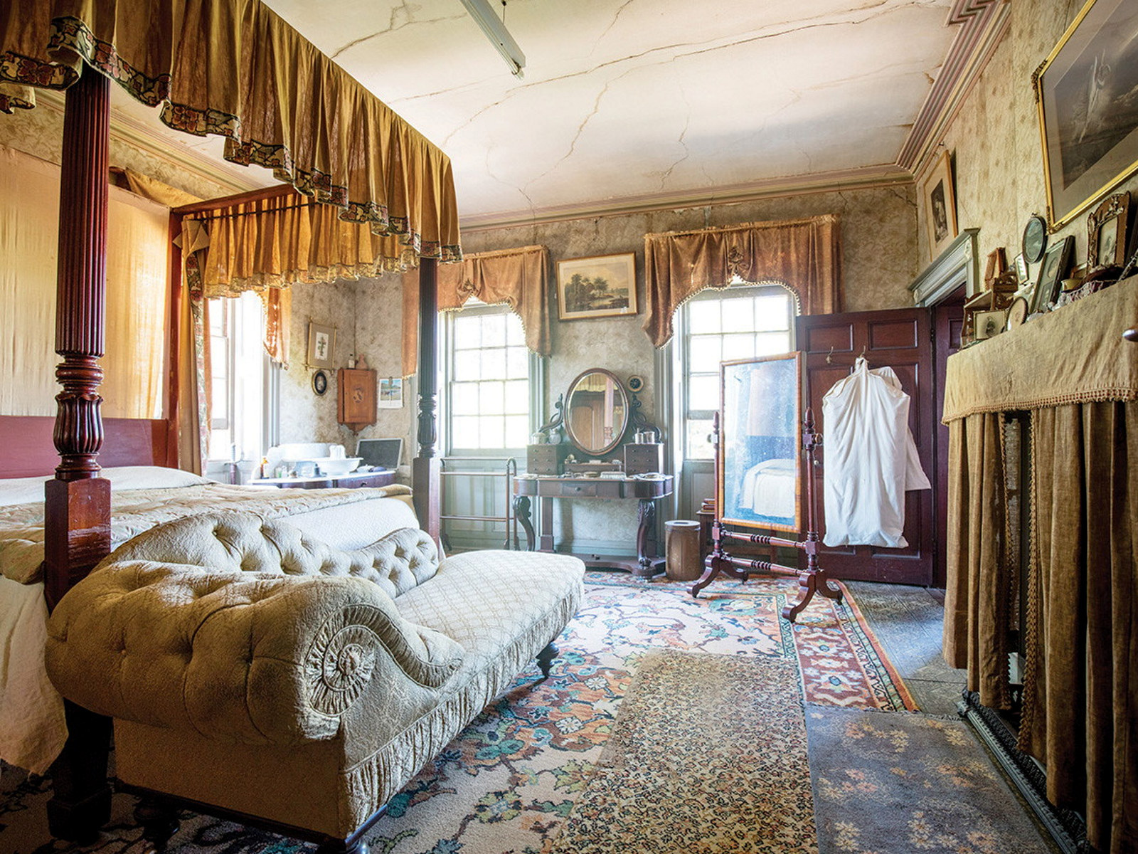 Ornately furnished bedroom with canopied bed and chaise longue to left and windows behind.