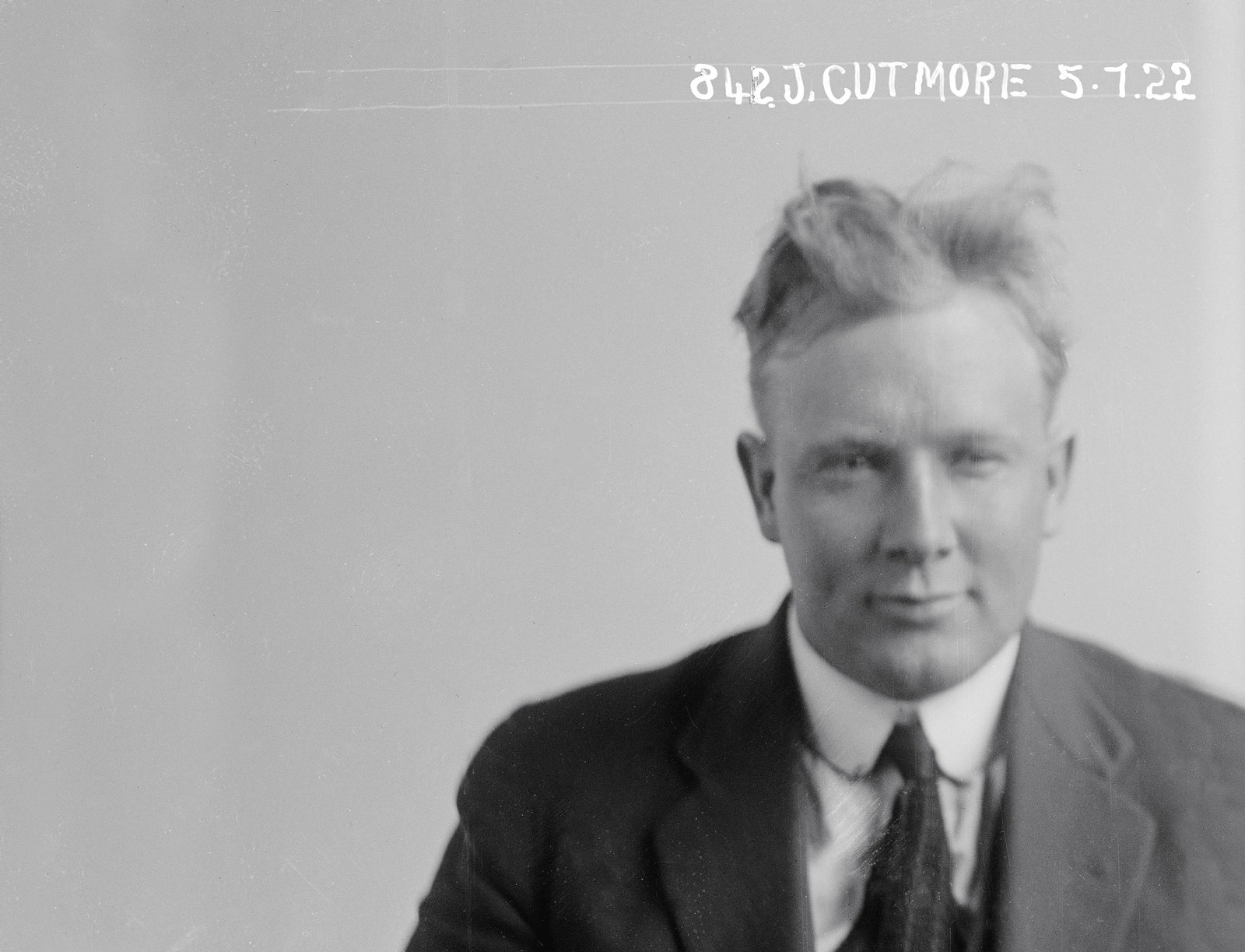 Black and white photo of a blonde man in a suit with scruffy hair. He is smirking.
