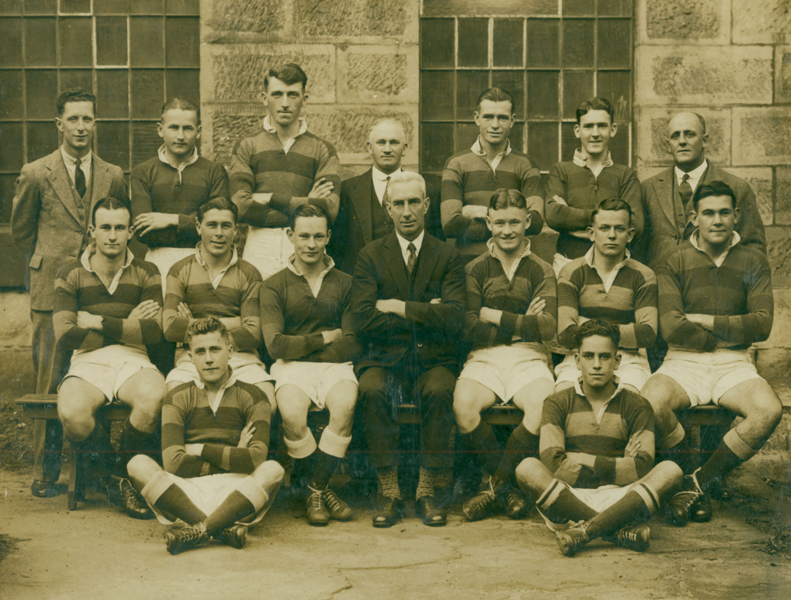 Group portrait of men's football team, players wearing striped jerseys, boots, socks and white shorts. There is a signle suited man in the centre of the group. A stone wall with large multi panelled windows is at the rear.