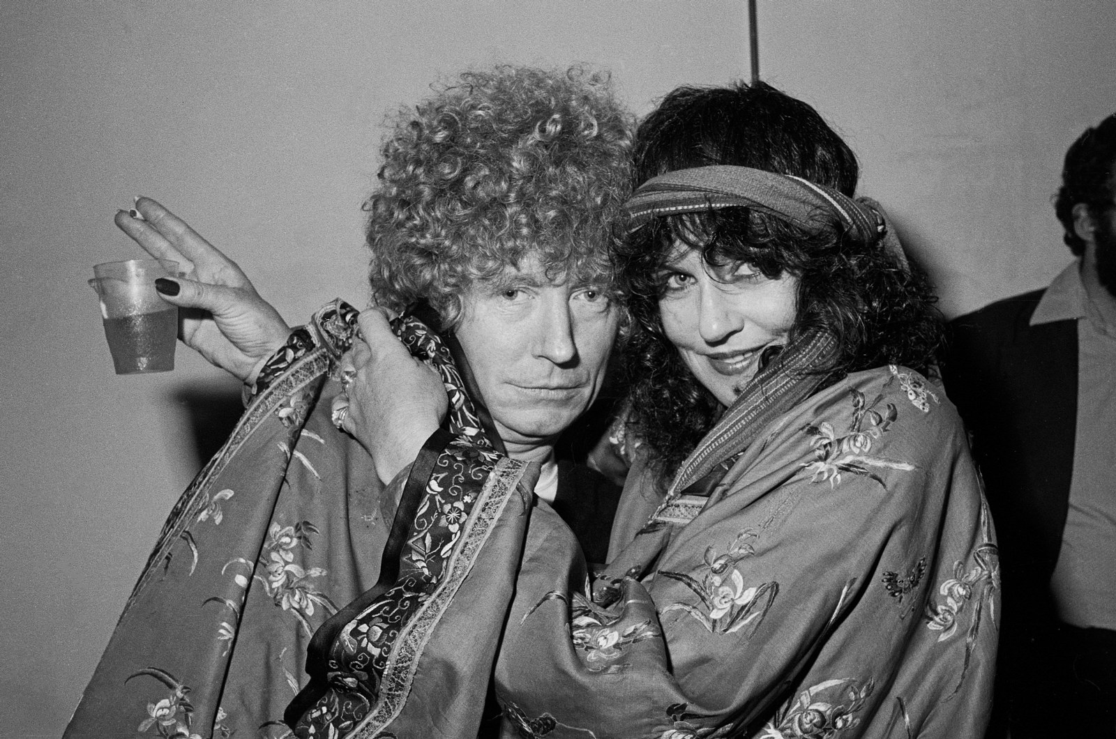 Man with curly hair being hugged by woman with headscarf holding drink and cigarette.