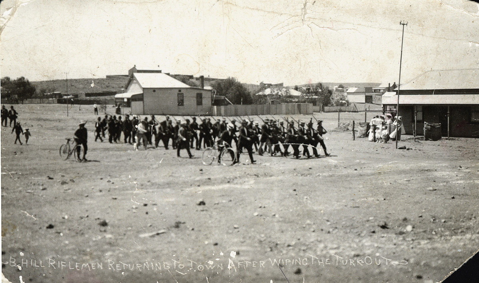 Group of men in formation in middle ground of large open area with buildings behind.