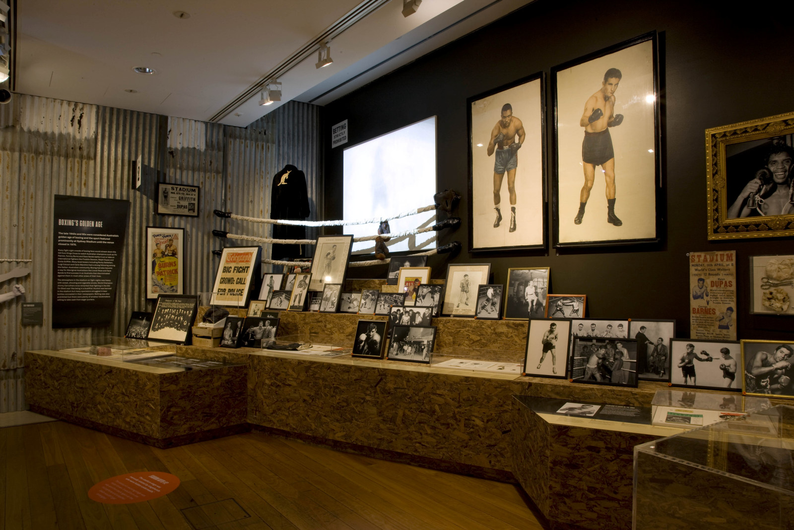 Exhibition interior showing framed photographs of boxers, posters and a screen on the wall.