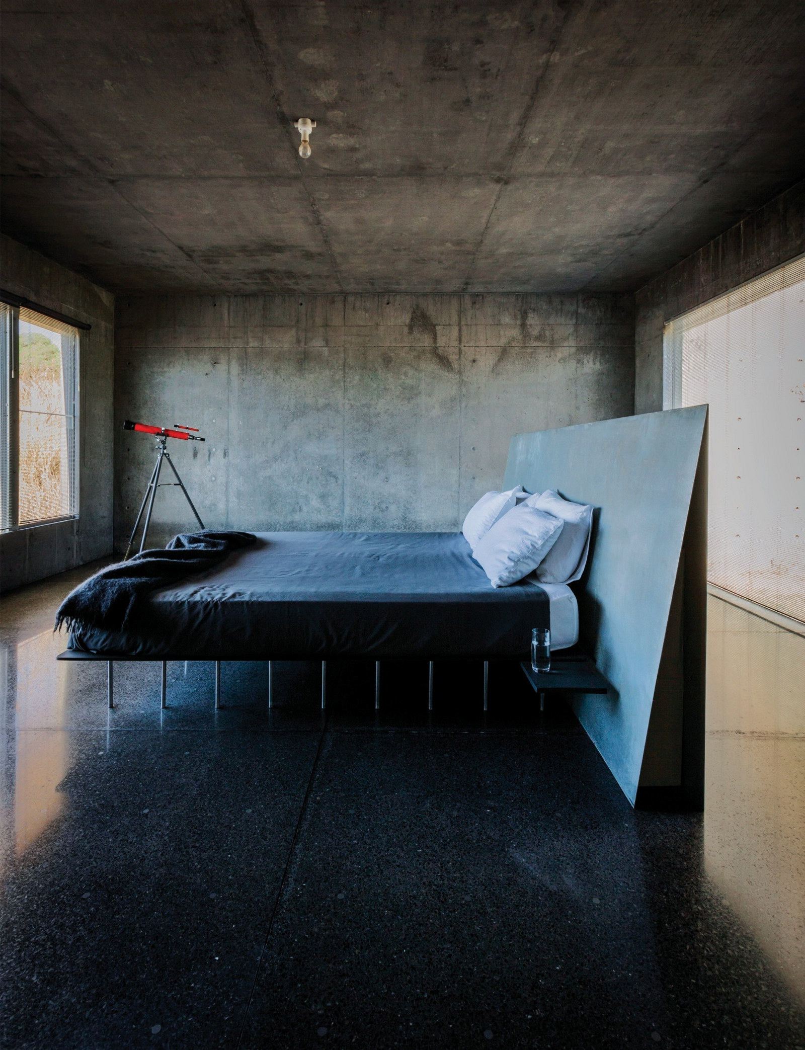 This is a photograph of a bedroom with a polished concrete floor with a concrete ceiling and large windows facing the double bed in the centre of the room
