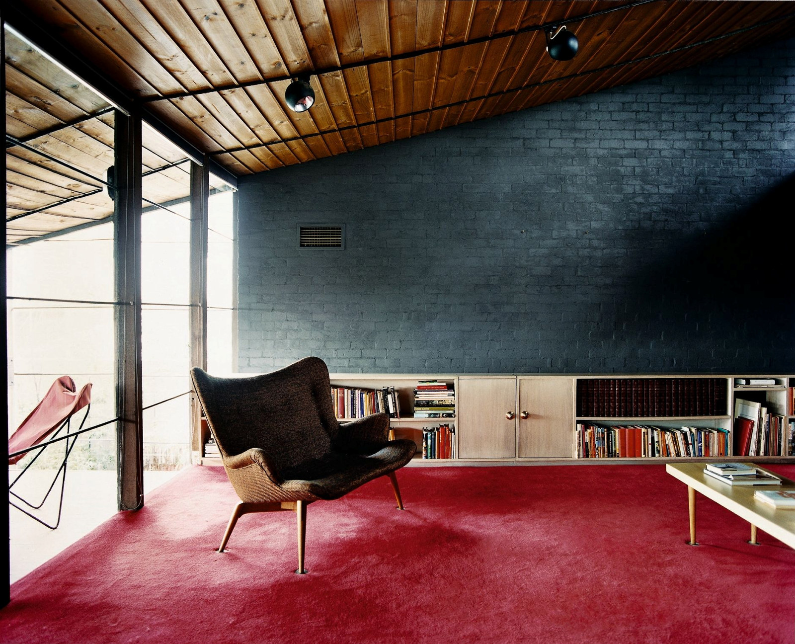 This is a colour photograph of a two seater couch in an open living space with deep red carpet and grey painted brick walls