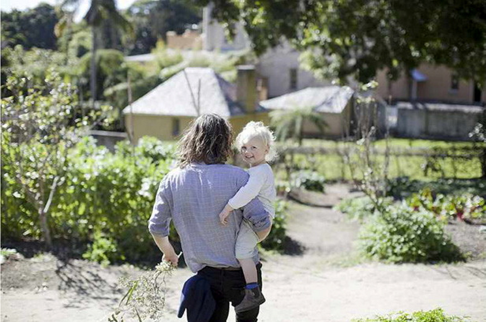 Man holding small child in garden facing away from camera with house in background.