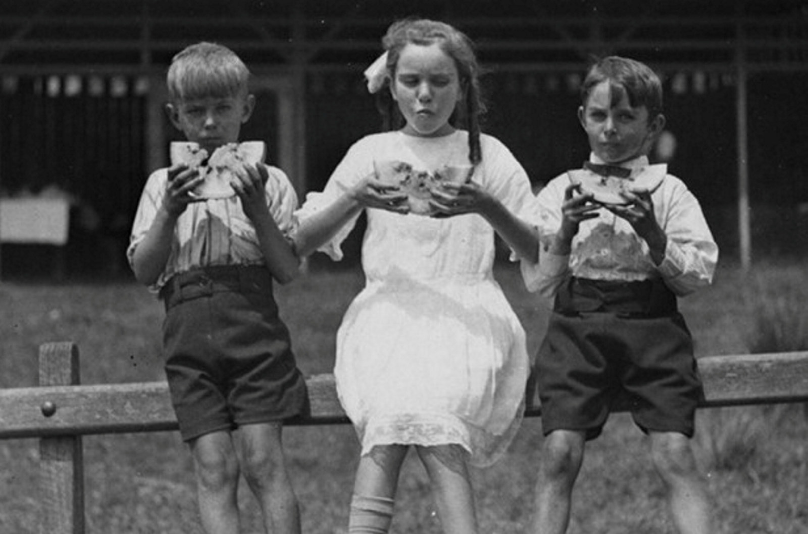 Black and white photo of three children sitting on a fence eating watermelon slices.