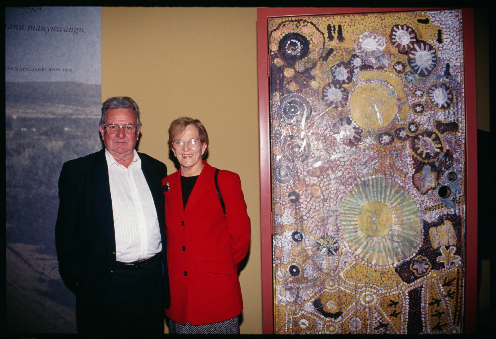 Man in black jacket and woman in red top in front of artwork.