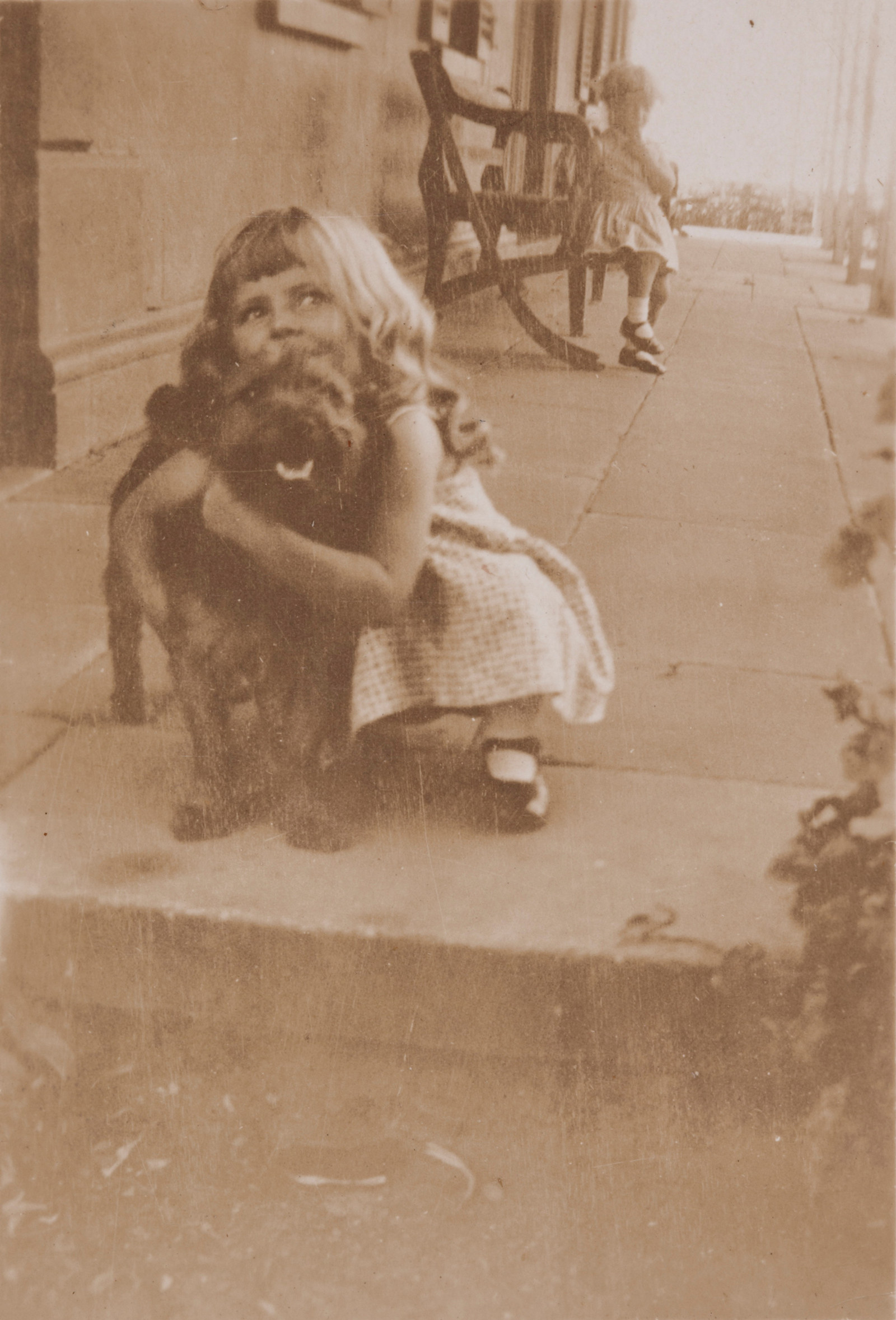 Sepia toned photo of young girl hugging dog on stone step with another young girl in background seated.