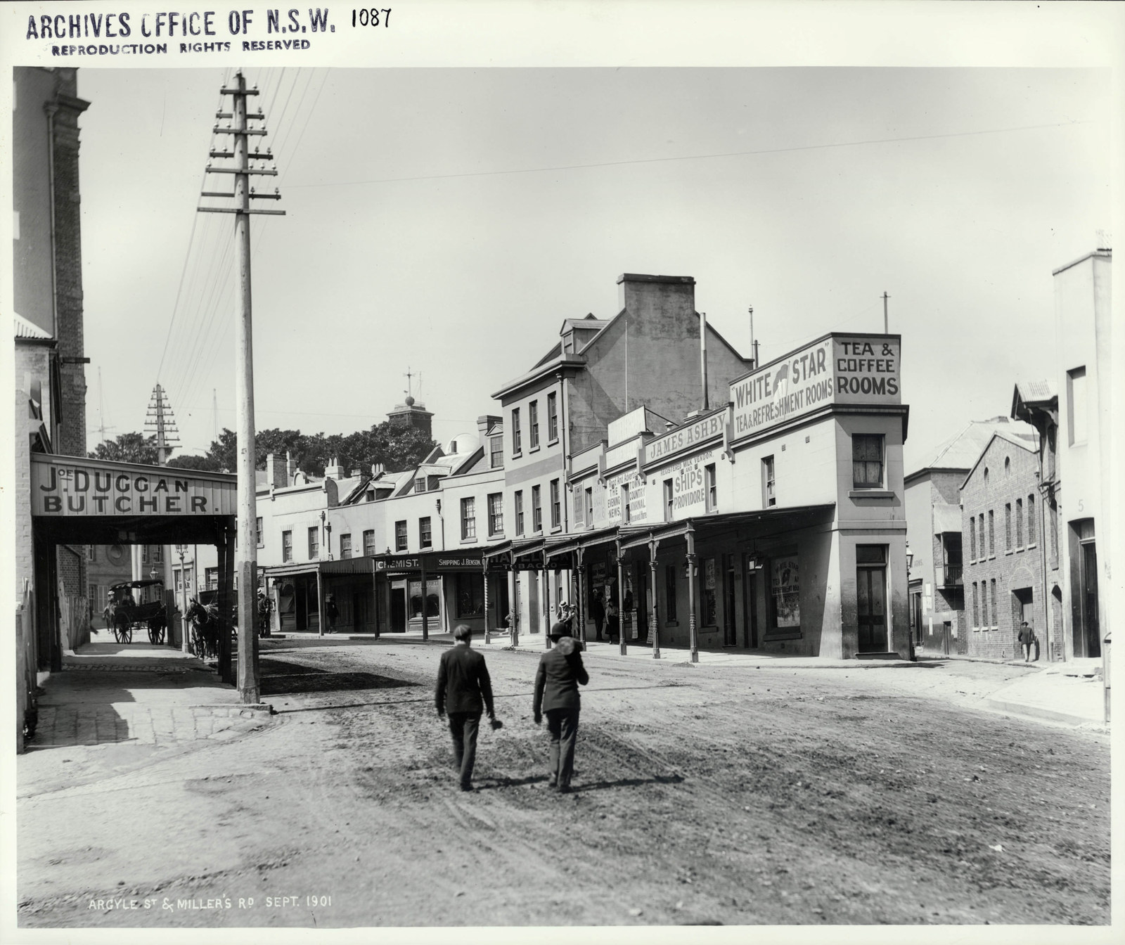 Photograph of street scene showing a row of two storey terraces. The street is lined by awnings indicating shops. Two men walk down the centre of the wide street.