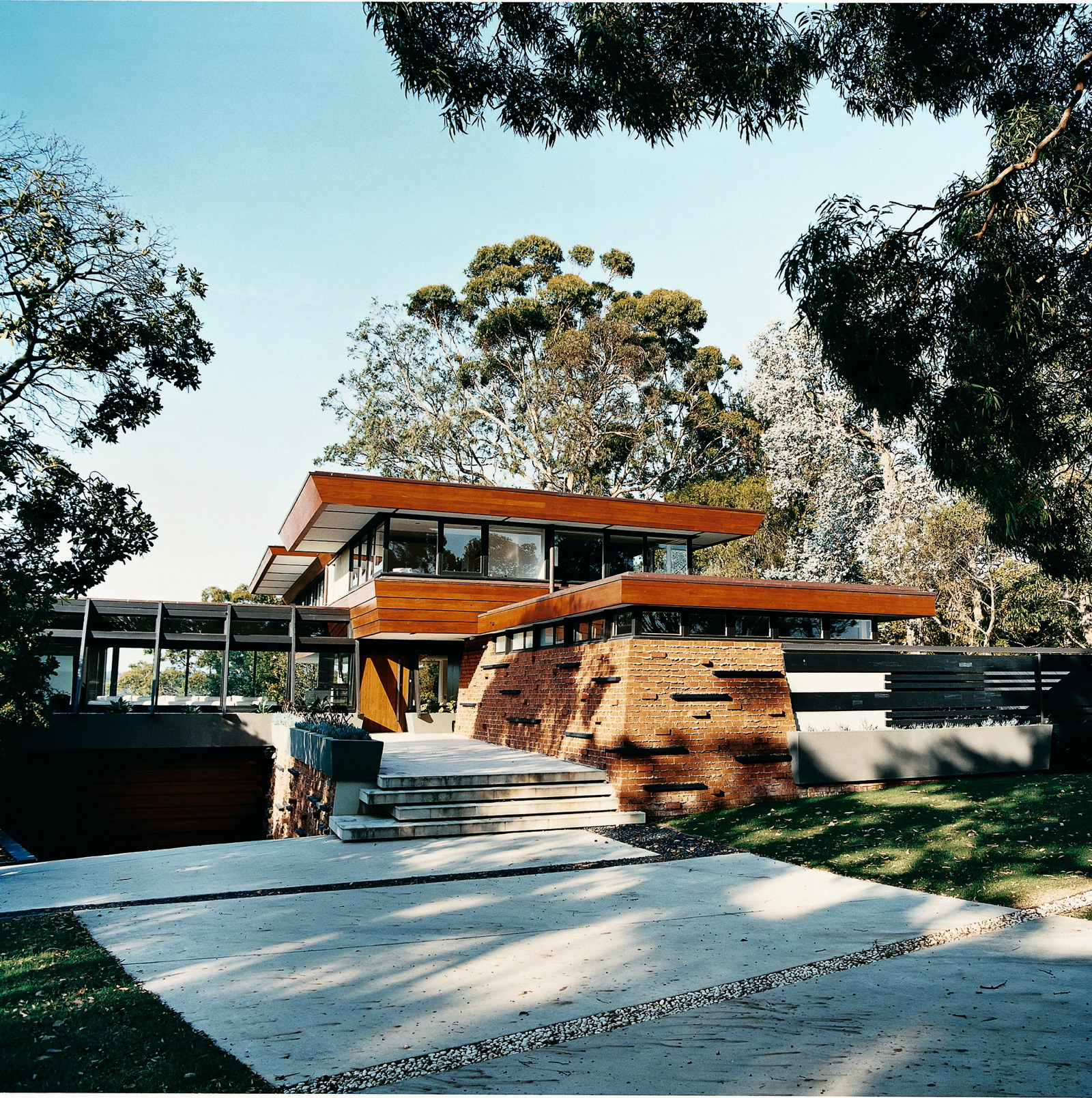 This is a front three-quarter photograph of a modernist 1950s house with a timber, glass and red-brick frontage and 5 low concrete stairs leading up to the front door