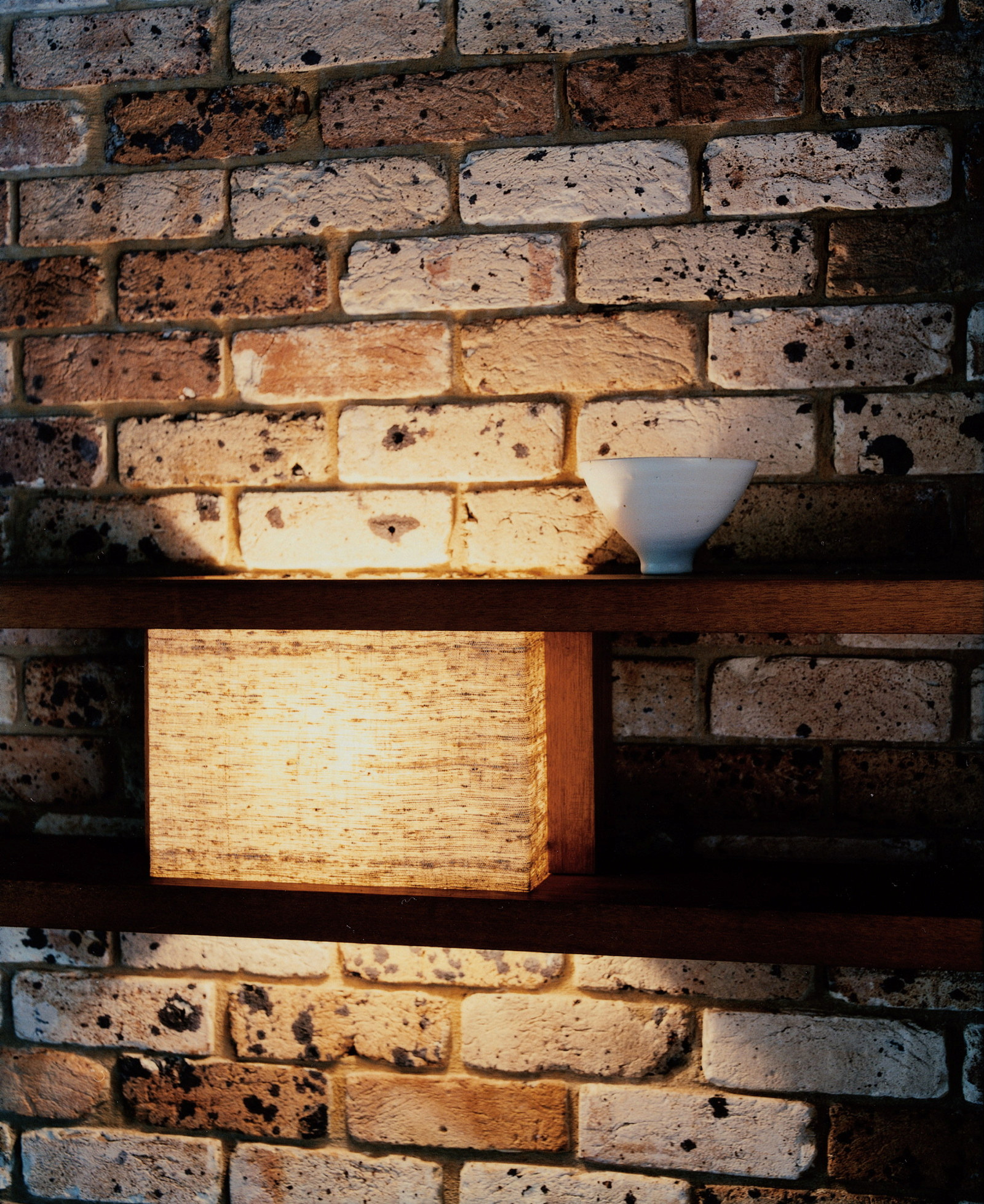 This is a detail photograph of a brick wall with two timber shelves. In one section between the two shelves a light has been created using a piece of linen.