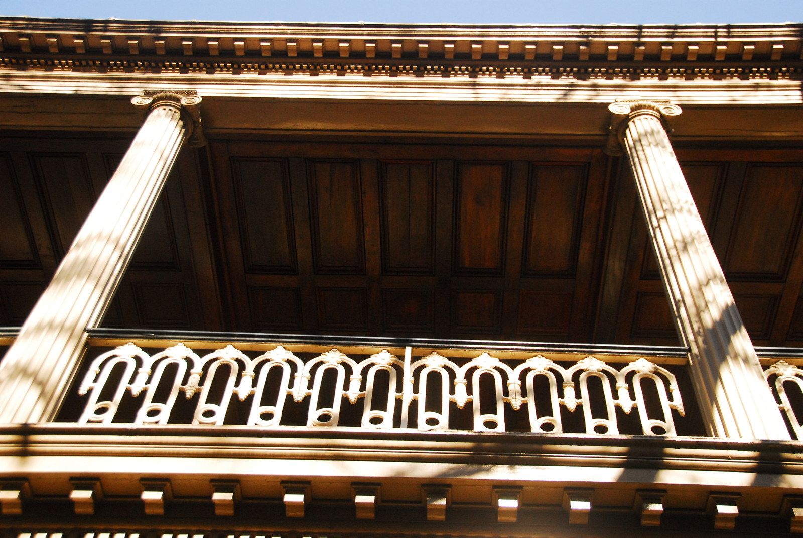 Looking up at darkened verandah, with columns, ironwork and two columns highlighted by sunlight.