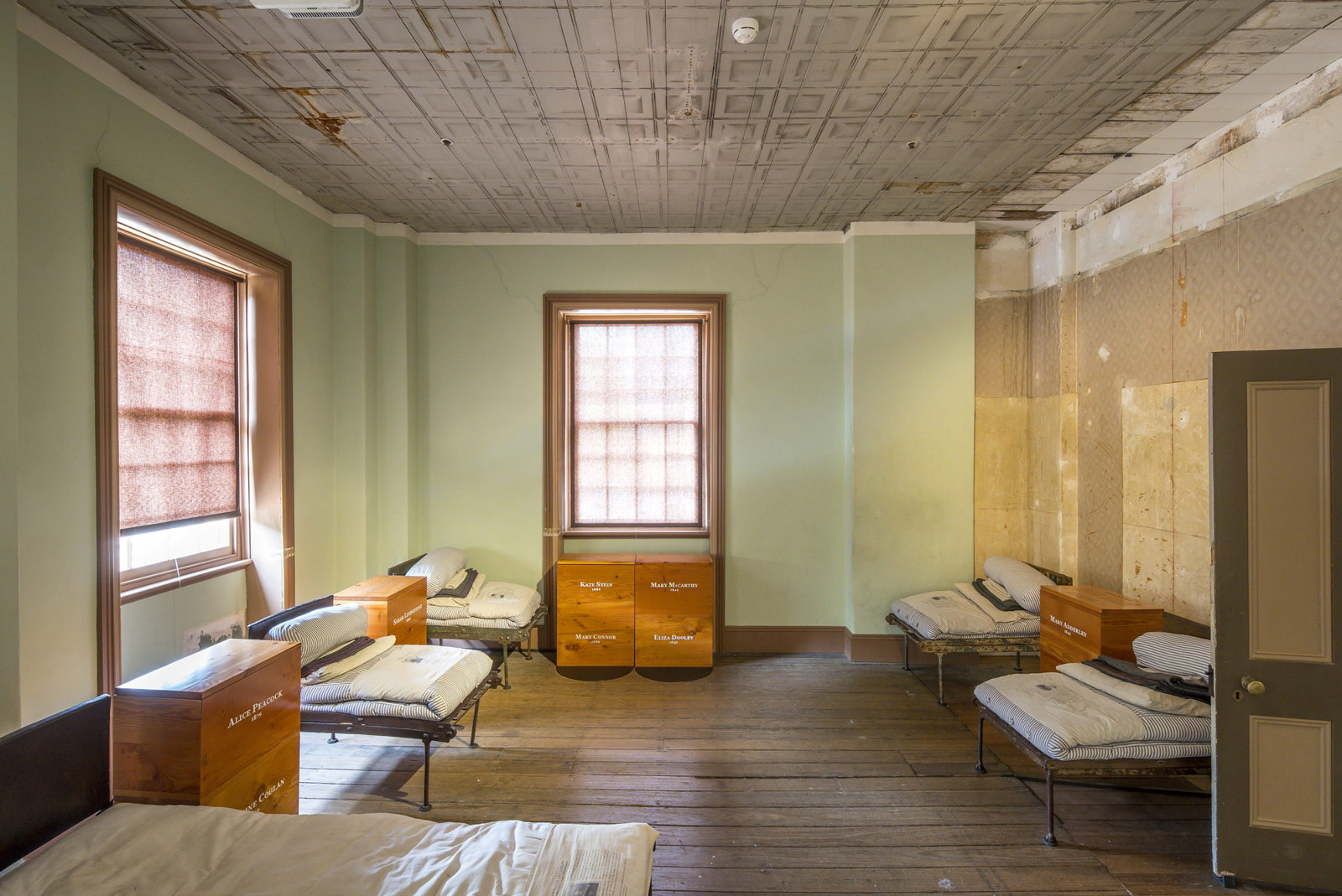 Room with wooden floor, green walls and windows to left, with simple beds made up to represent dormitory.