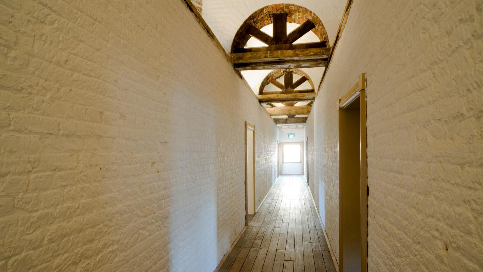 Long corridor with wooden floor, painted brick walls and wooden arches above, leading to backlit window at far end.