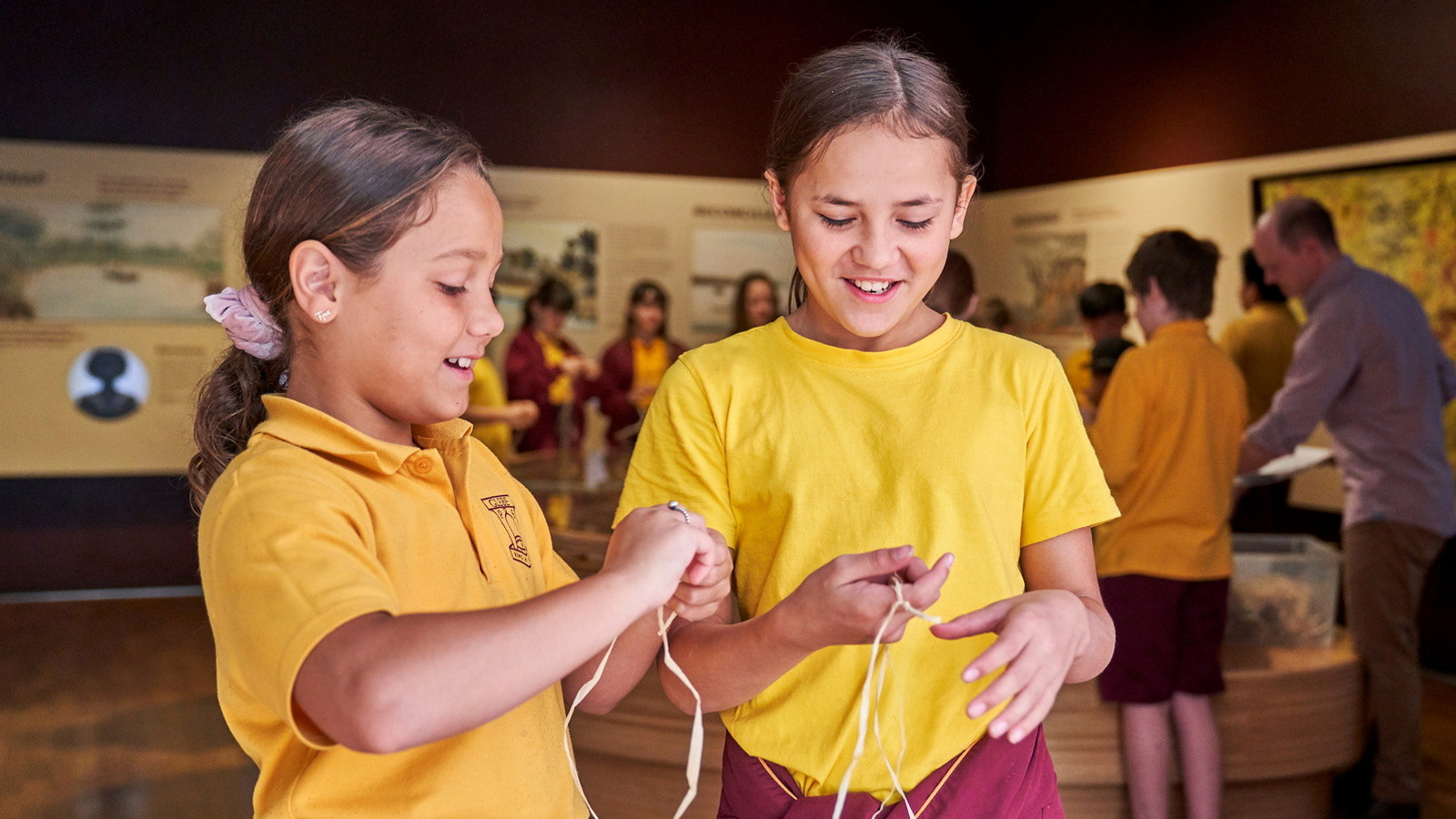 Two girls in school uniform making string in exhibition space with other students behind.
