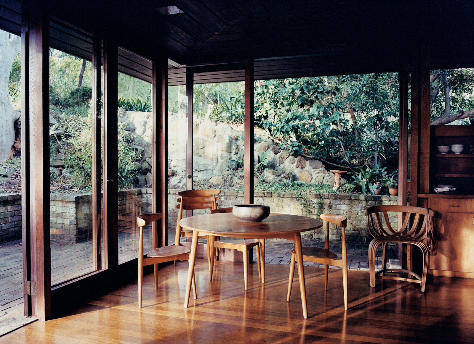This is a photograph of a timber floored room open with large glass sheets on two walls and a timber dining table and chairs