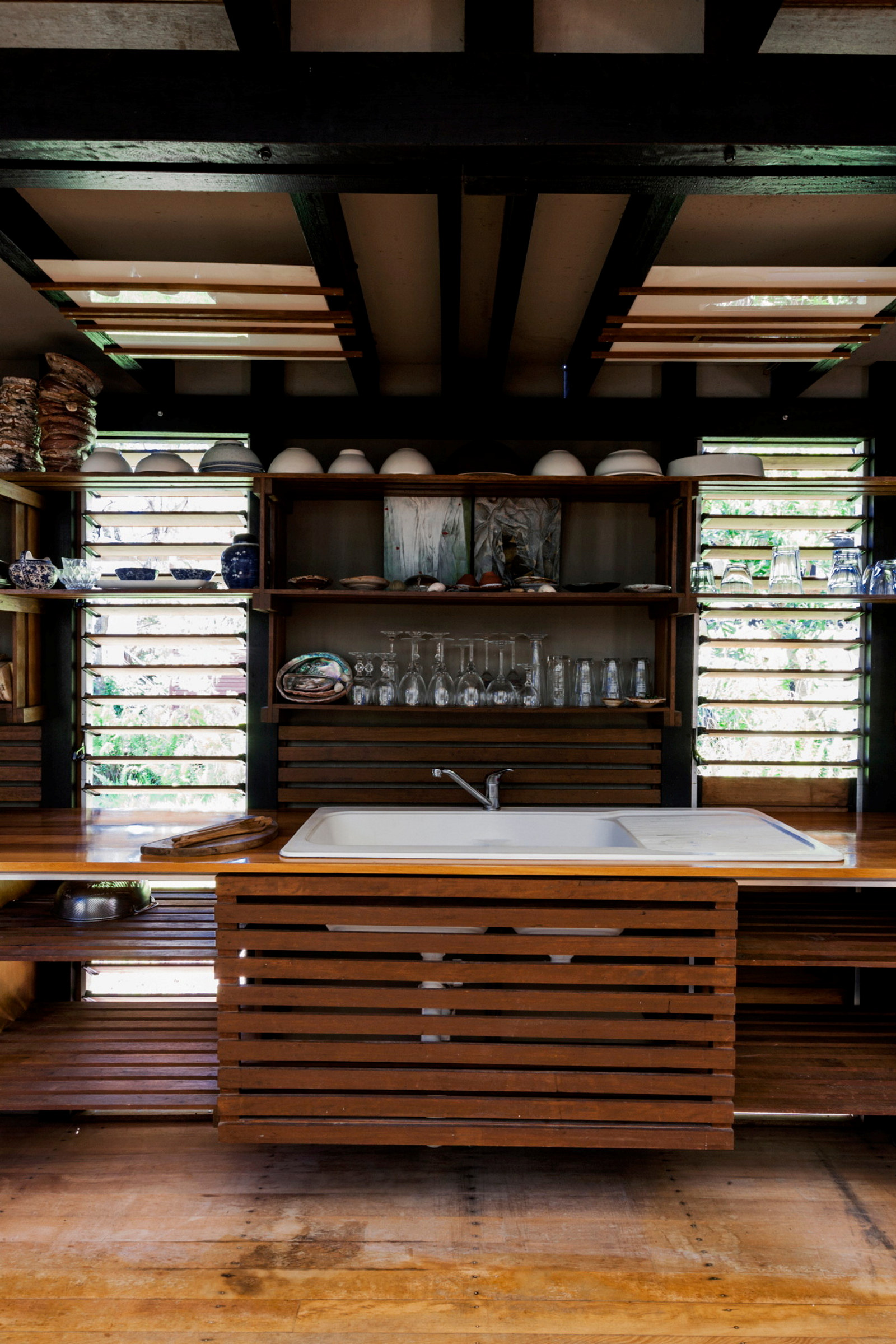 This is a photograph of a timber and glass clad space with a large white ceramic sink in the centre and bowls, plates and glasses on the back wall