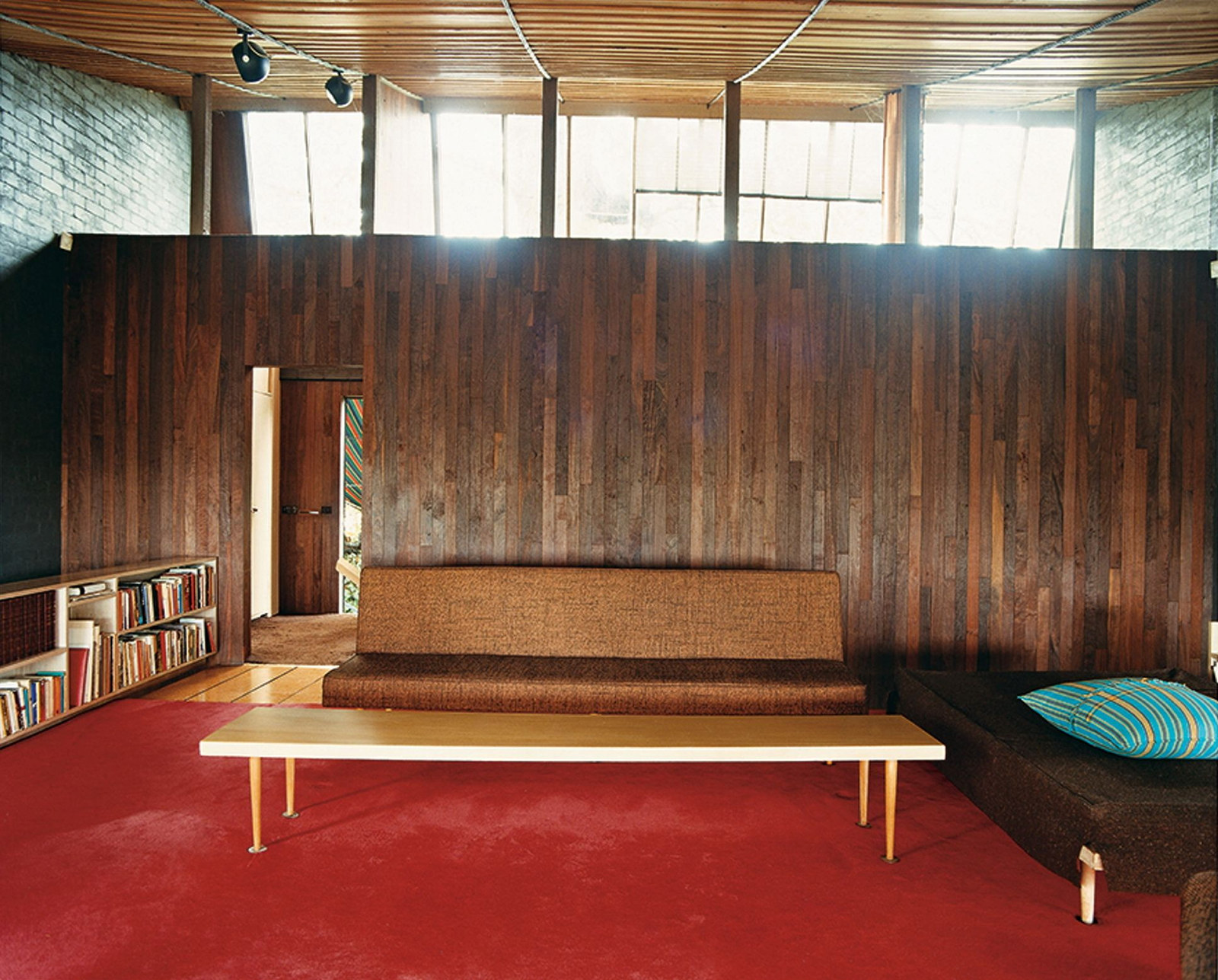 This is a colour photograph of a simple 1950s style sofa with a low timber coffee table in front with a deep red carpet floor and timber wall behind