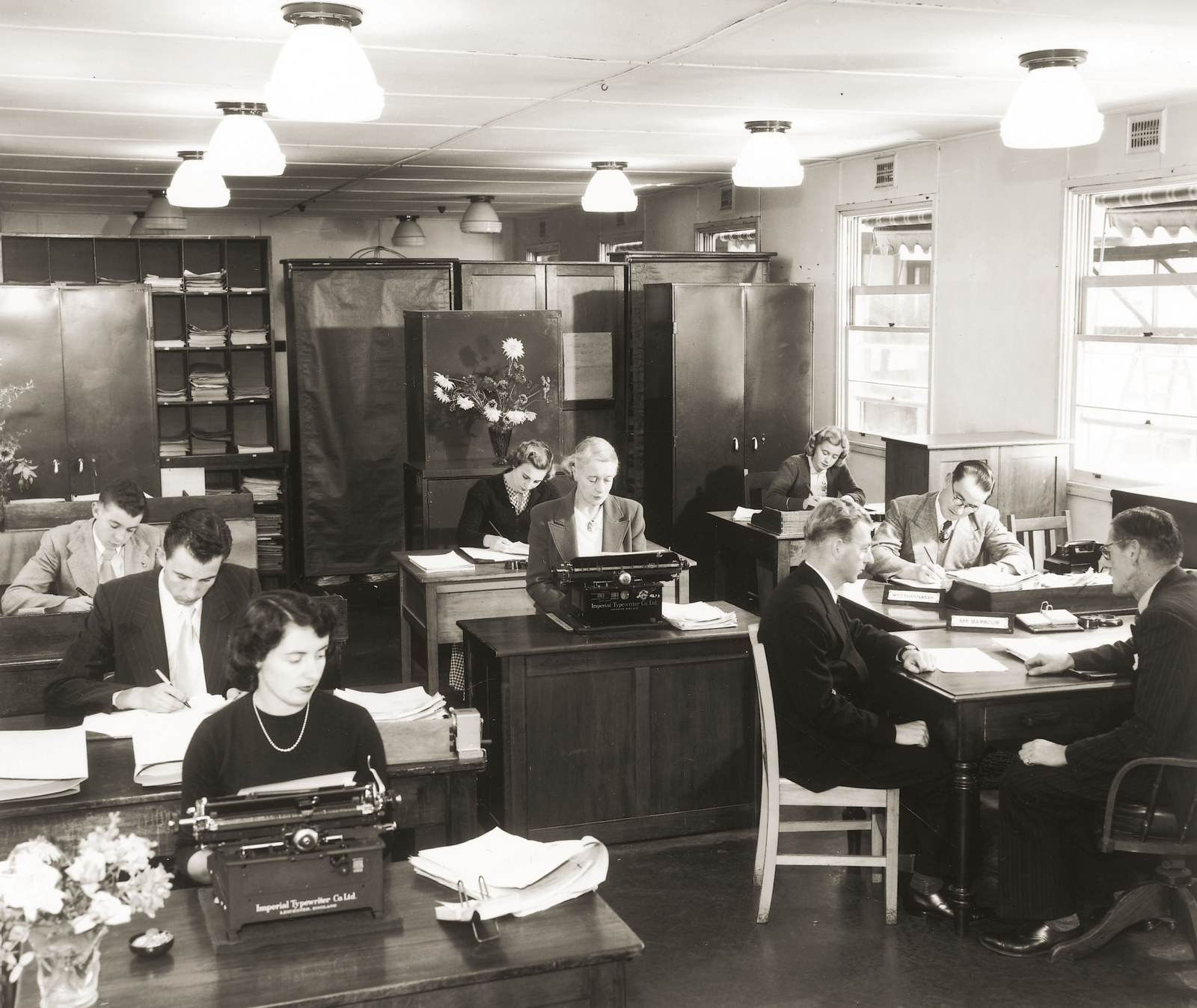 Office with several women on typewriters and men working at desks on paperwork. The room is filled with heavy office furniture, low ceiling and rows of lights.
