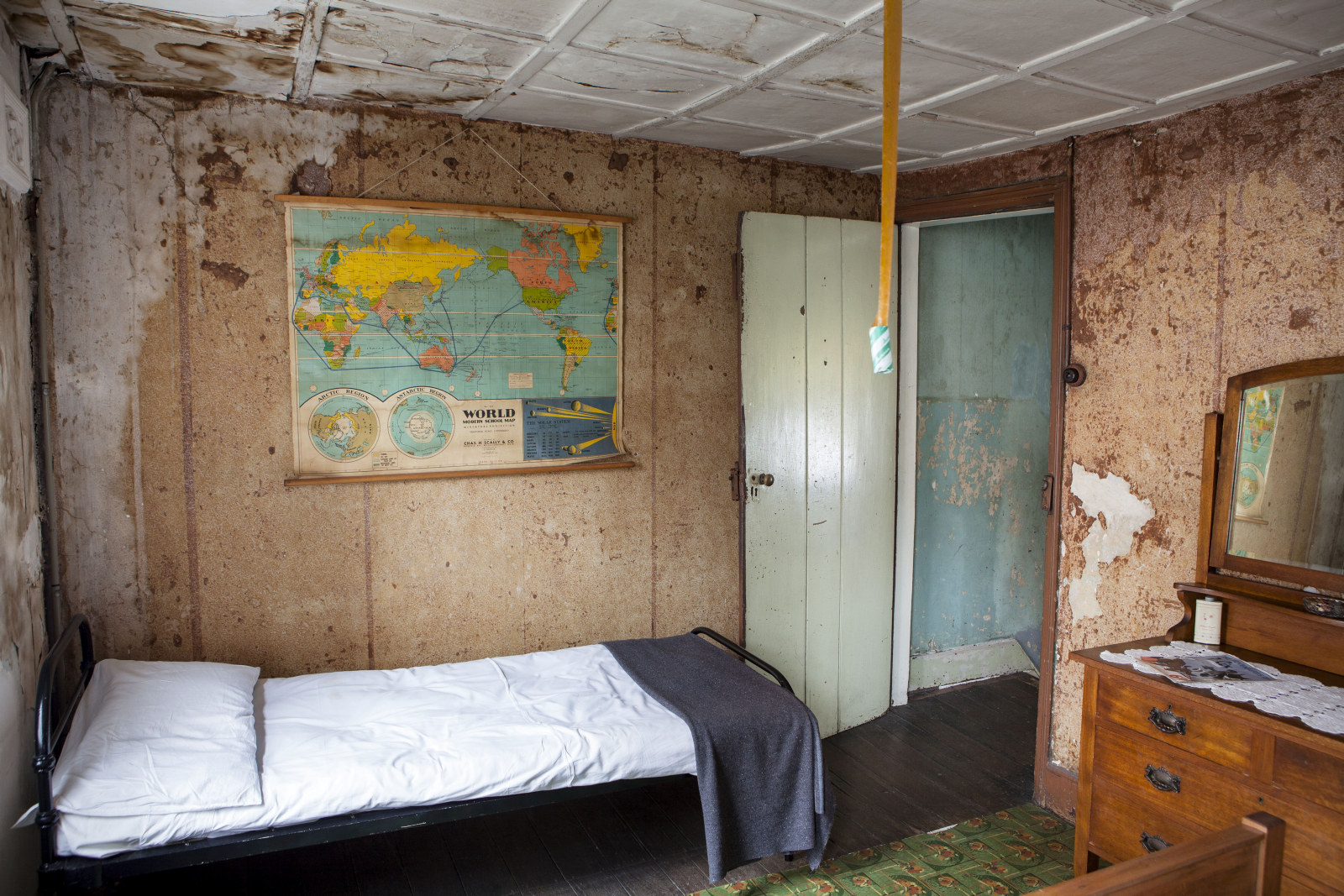 View of a bedroom with faded and torn wallpaper with an old map of the world on one wall, a single bed, dressing table and view through a door to the top of a staircase, with faded and damaged paintwork.