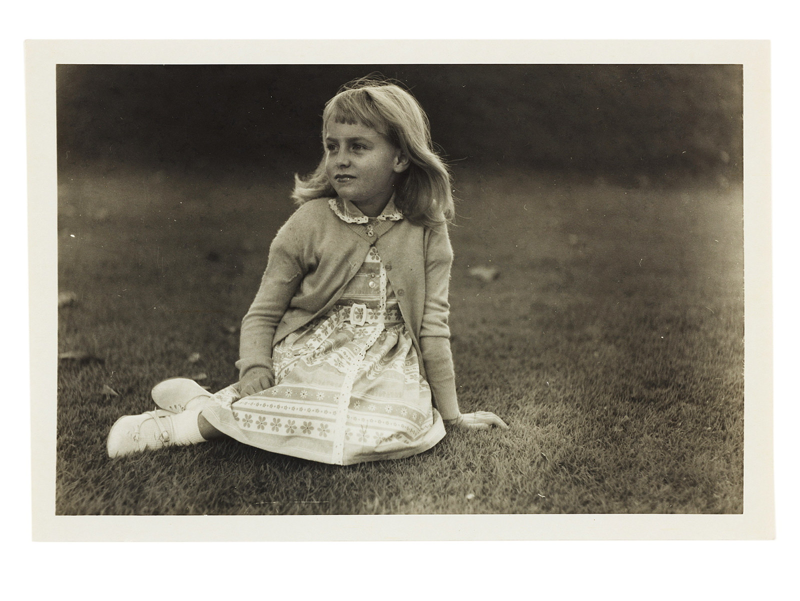 Black and white photo of young girl seated on ground with legs folder to her right.
