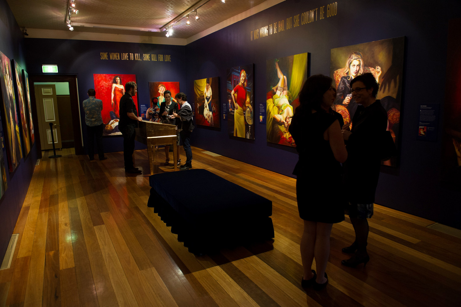 People milling around the main exhibition space during the opening of the exhibition. The room appears dimly lit with the paintings juming out.