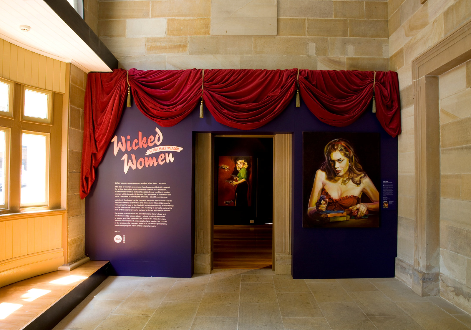 A red curtain hangs over the entranceway to the wicked woman exhibition. A text panel sits to the left of the door while a portrait of Tara Moss hangs to the right.