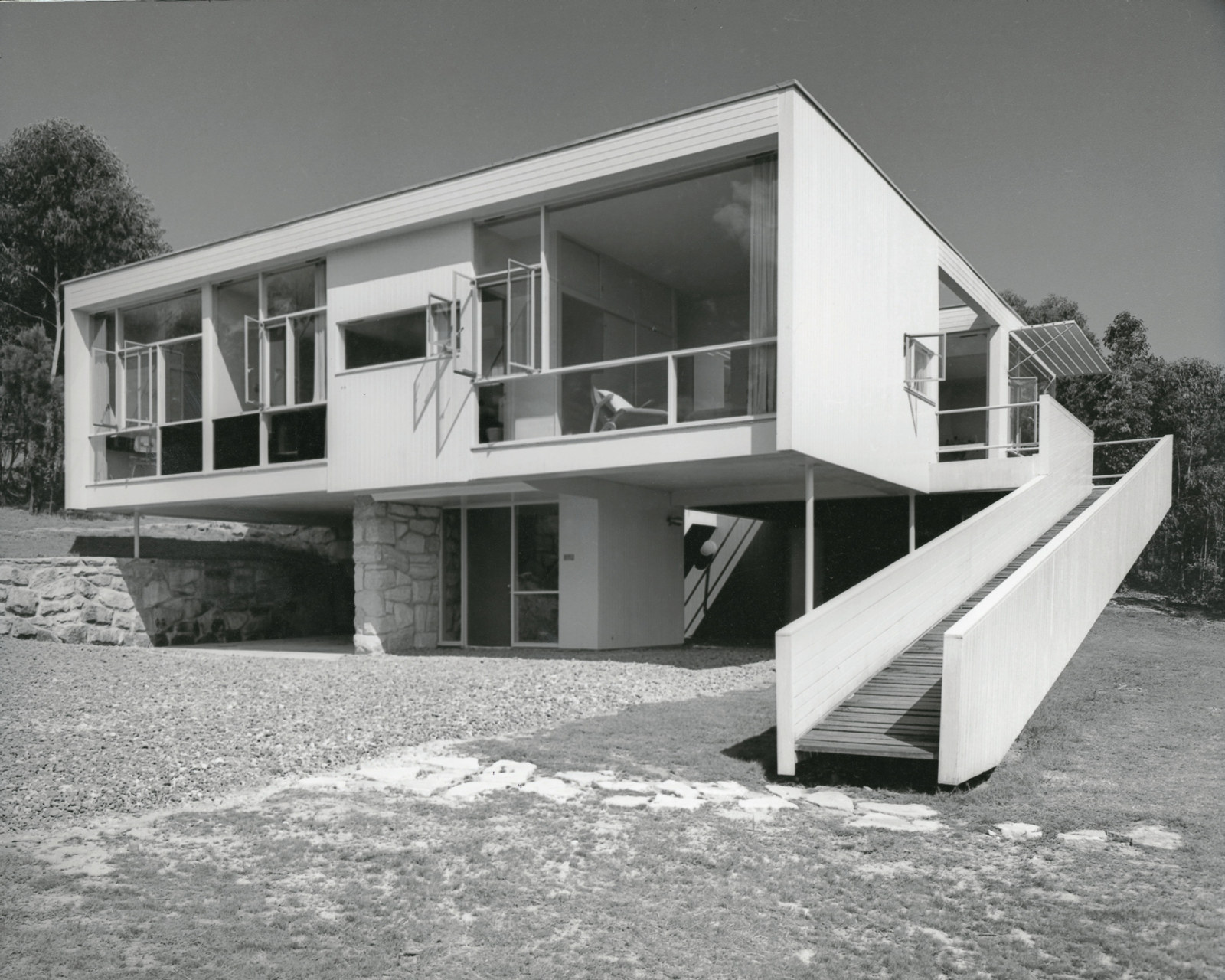 Diagonal B/W view of awesome white box-like house with large picture windws, suspended on poles and entrance lobby, with long sloping ramp connecting deck to ground and driveway area.