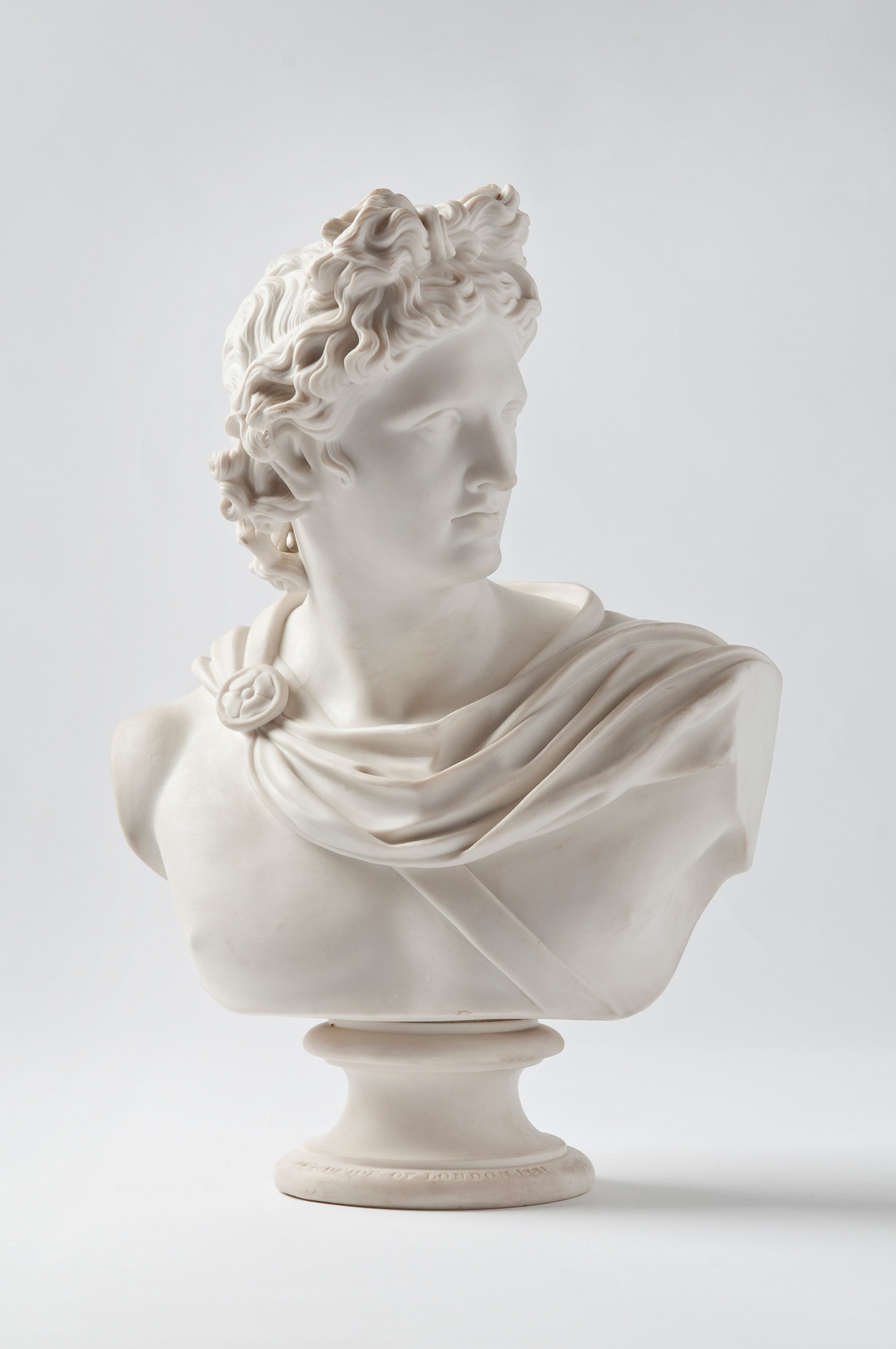 White bust of man wearing wreath in hair and drapery around neck, on pedestal.