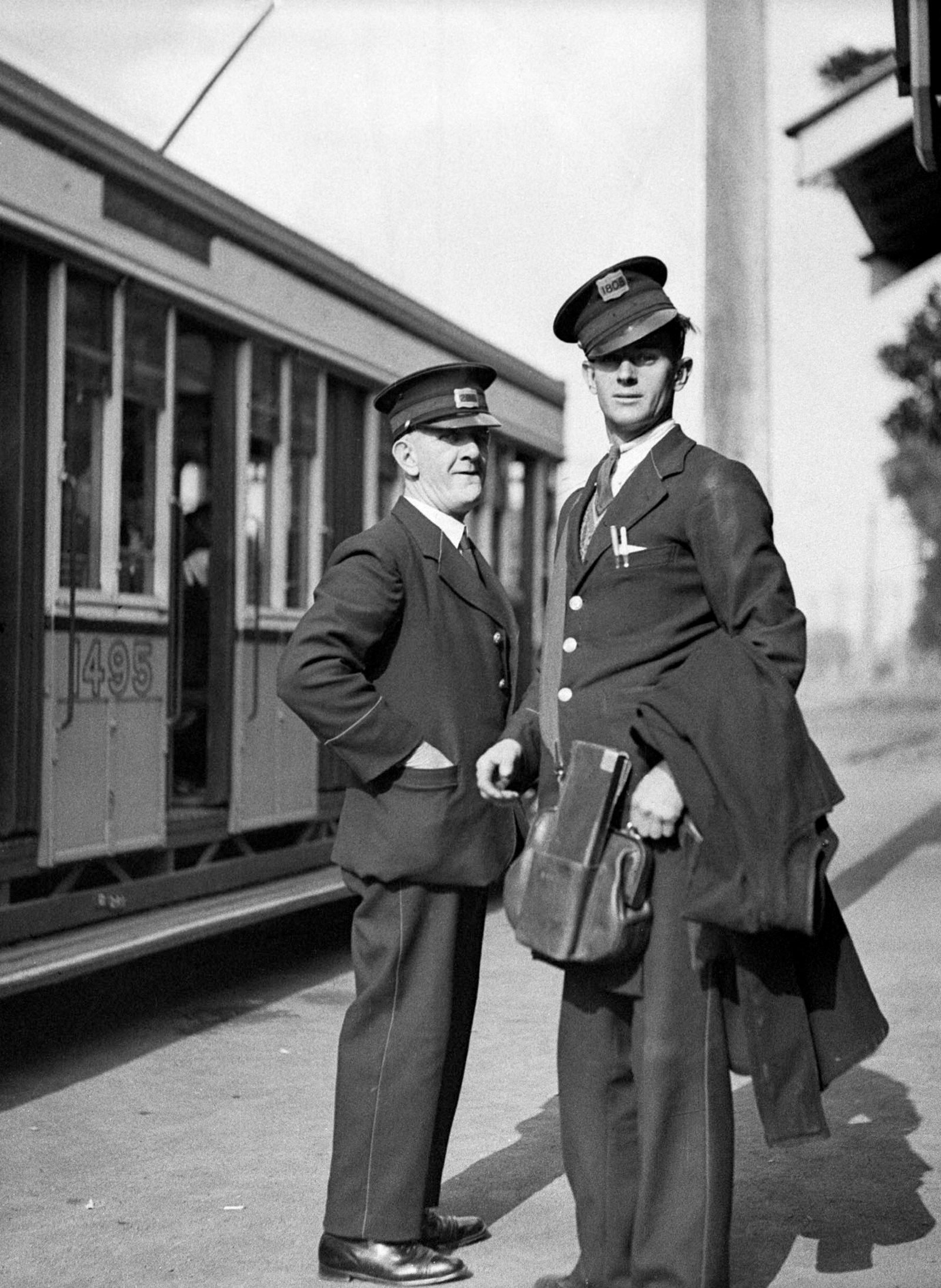 Two uniformed tram way men stand on the street beside a tram. One carries leather bag on his shoulder.