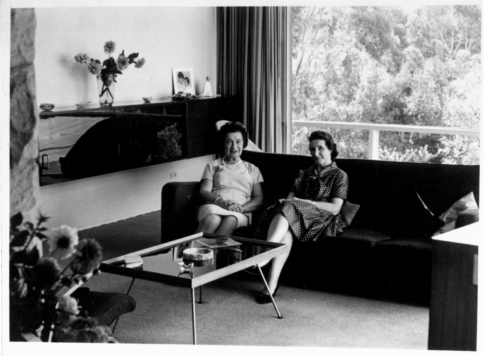 B/W photo showing two women sitting in couch in living room with coffee table in front, sideboard beside them and large window and curtains, with trees visible outside.