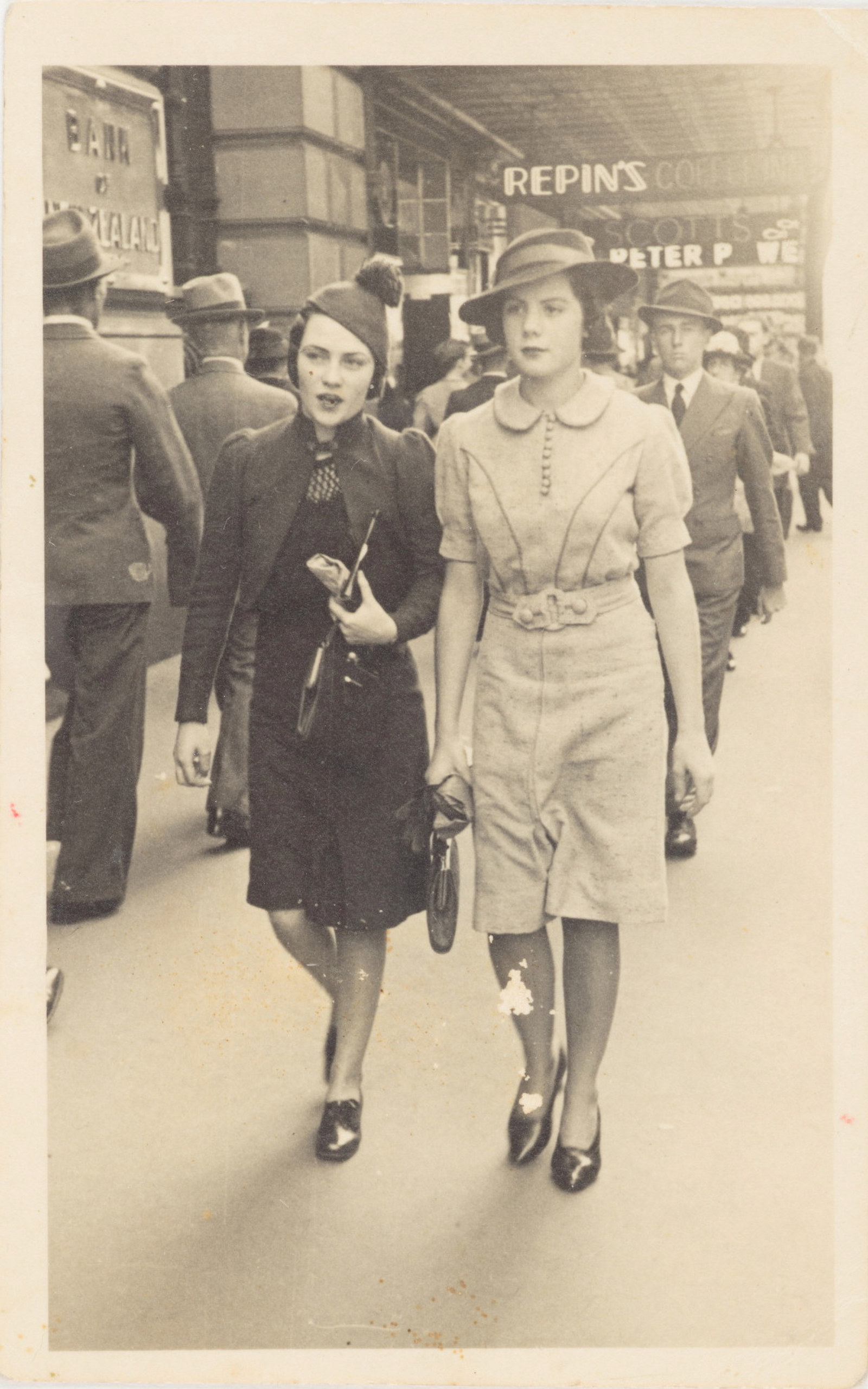 Sepia toned black and white photo of two women in hats walking down street.