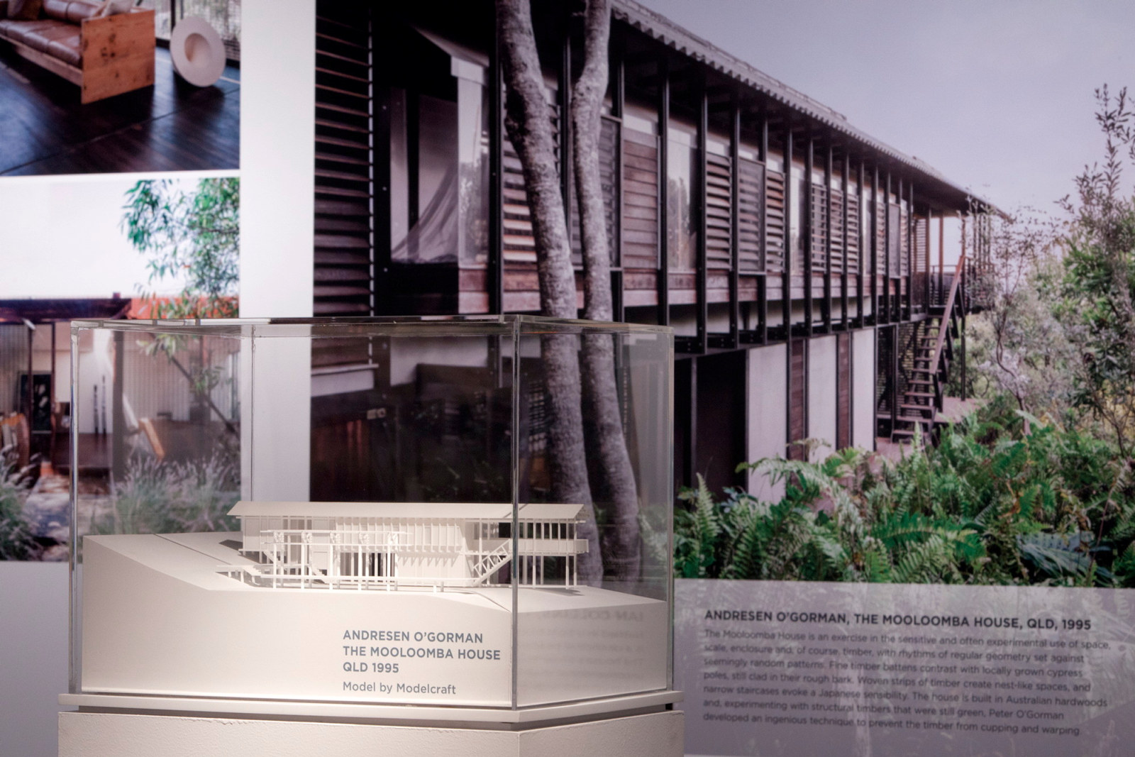 This is a photograph of a detailed architectural model in a case with a graphic panel of the house on the wall behind