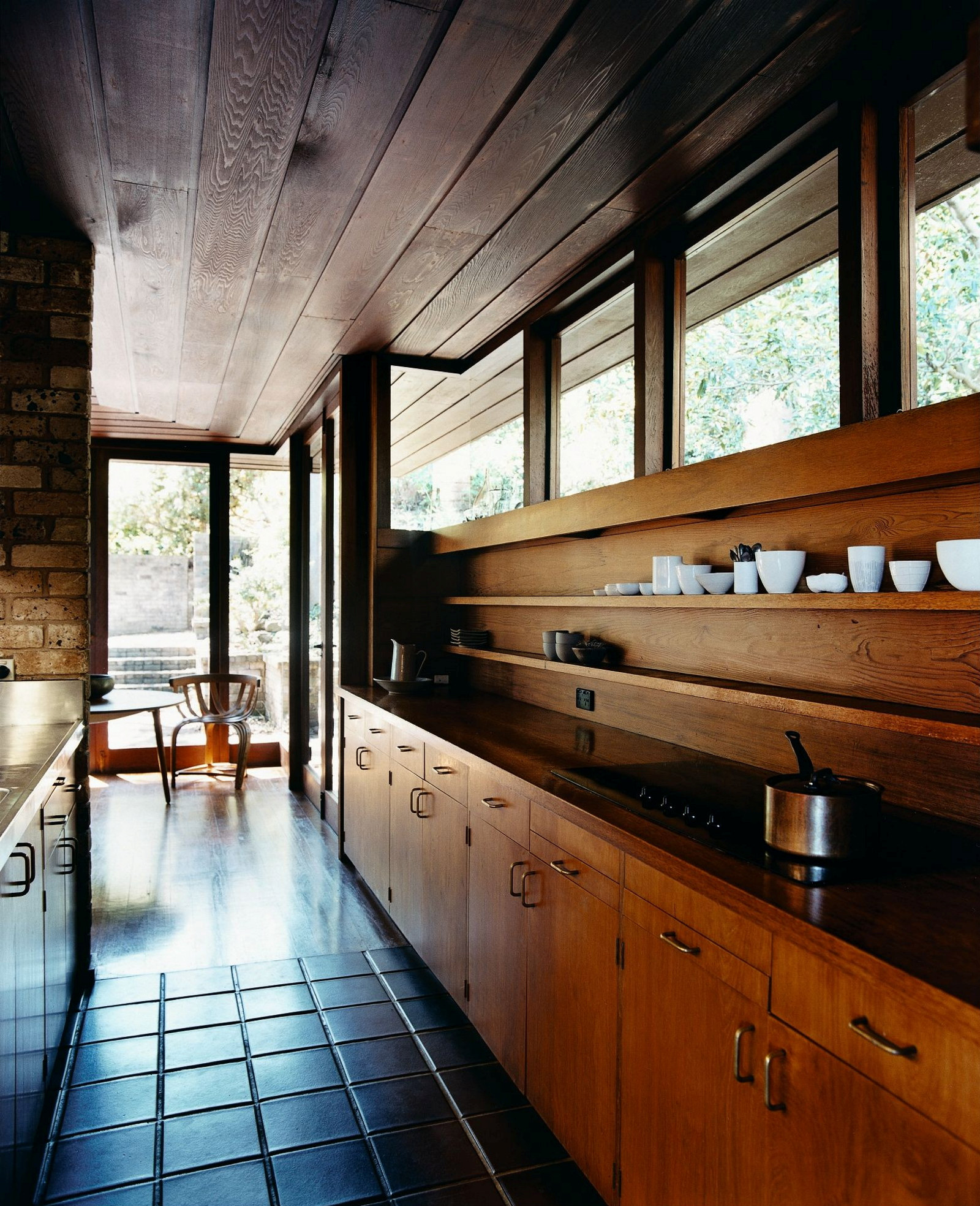 This is a photograph of a gallery style kitchen with a tiled floor. Timber is used for the ceiling, cupboards and benches.