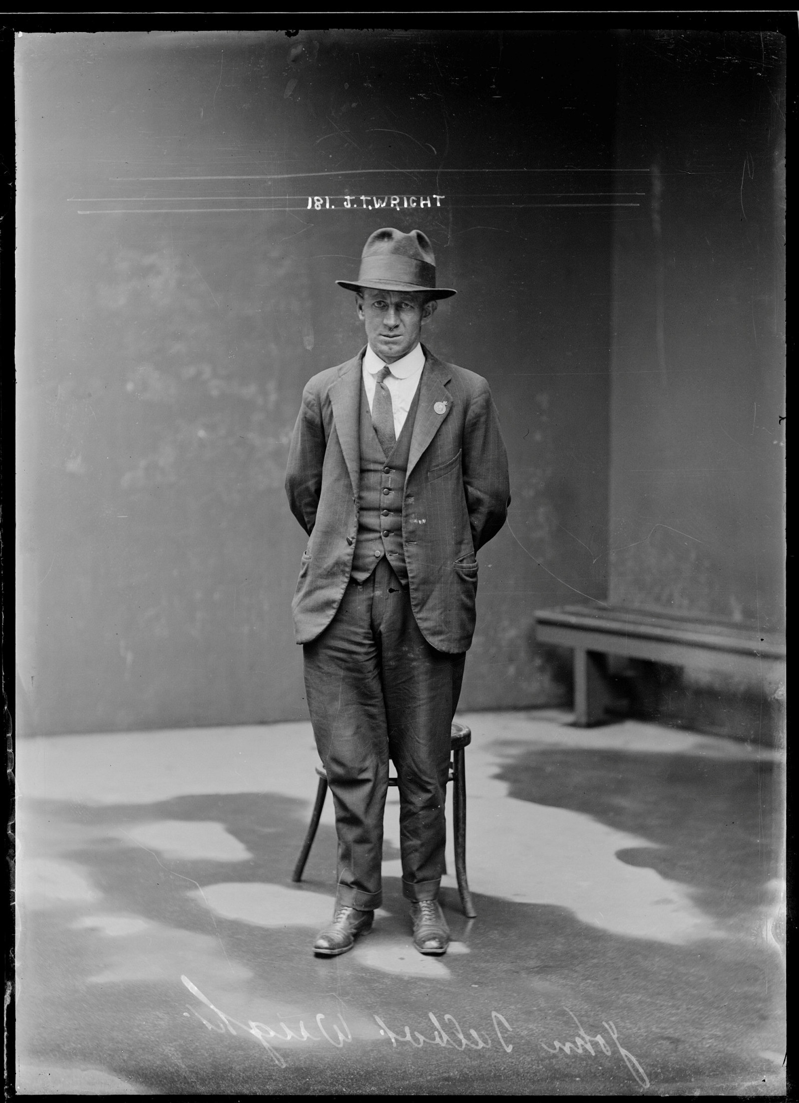 Full length police photo of man in suit and hat, standing in front of chair.