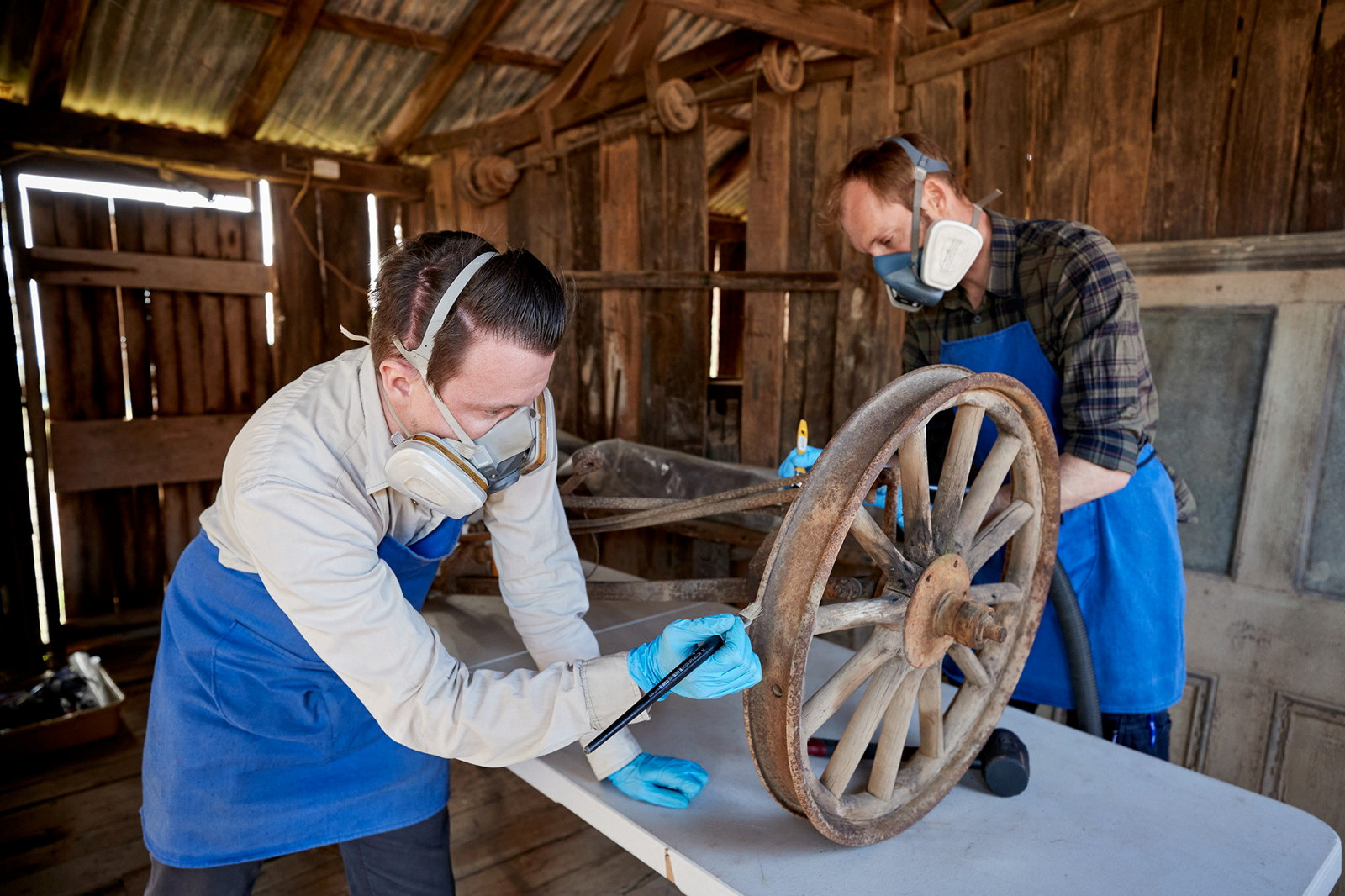 Two men wearing blue aprons and masks working on object inside wooden shed.