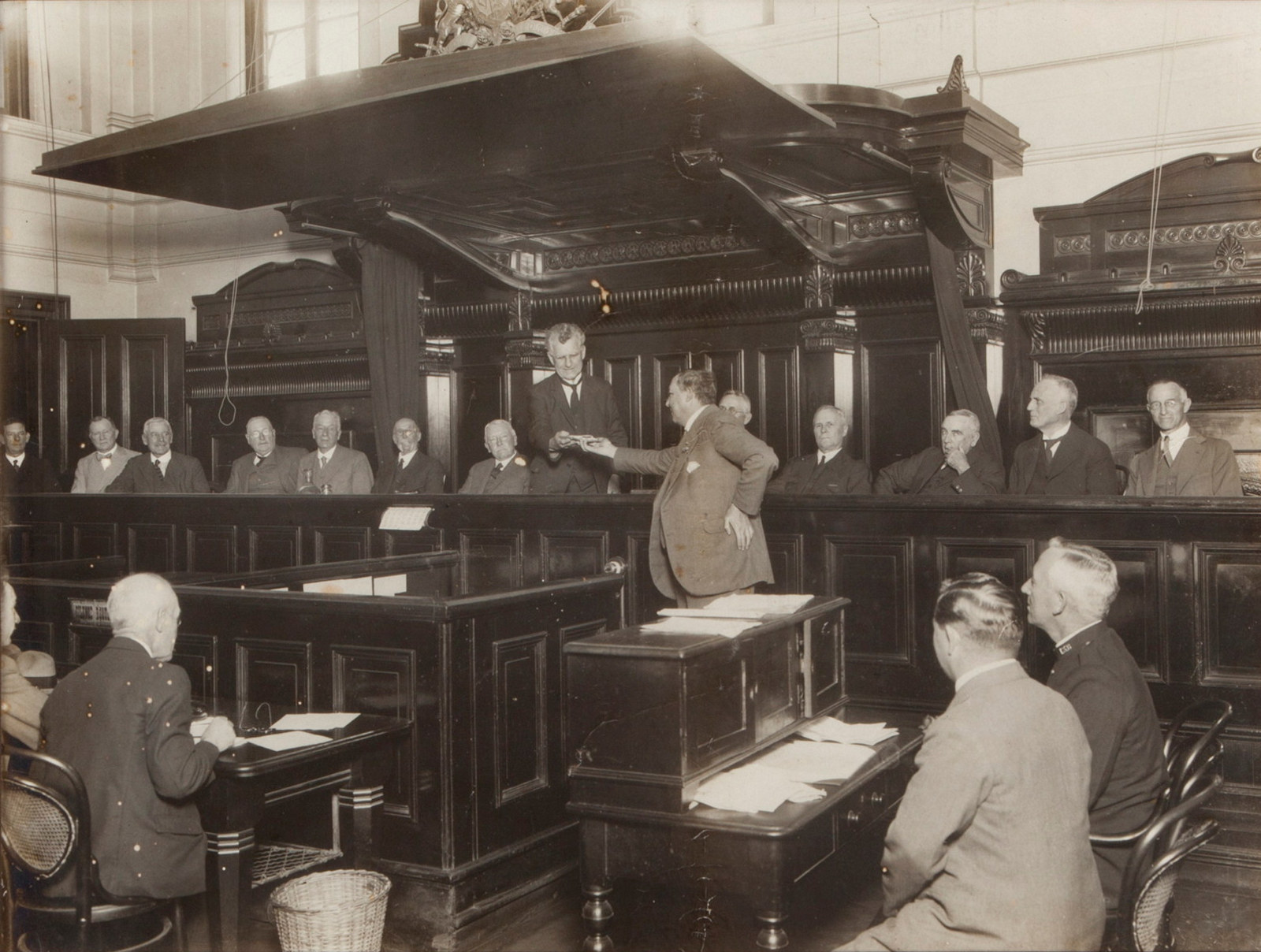 Courtroom full of men, focused on Magistrate in imposing wooden chair.