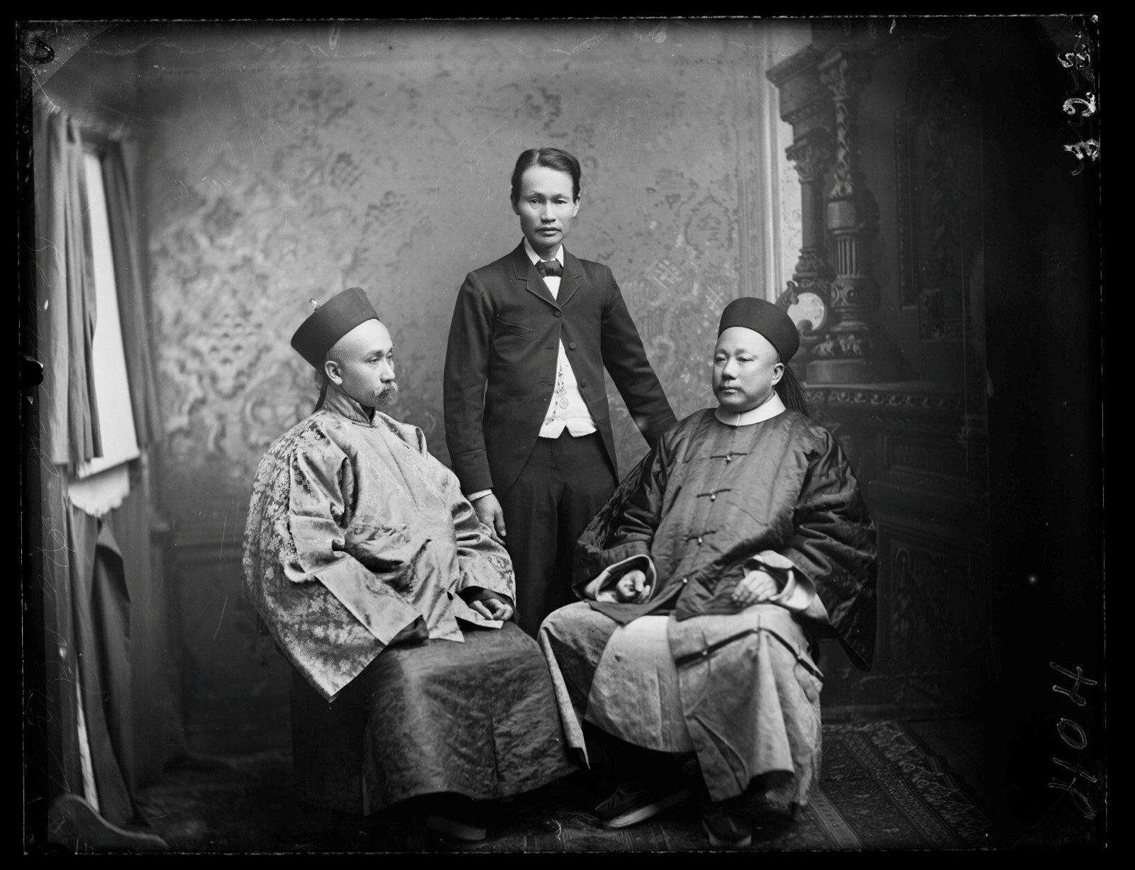 Black and white photograph of two seated men flanking a central man standing.