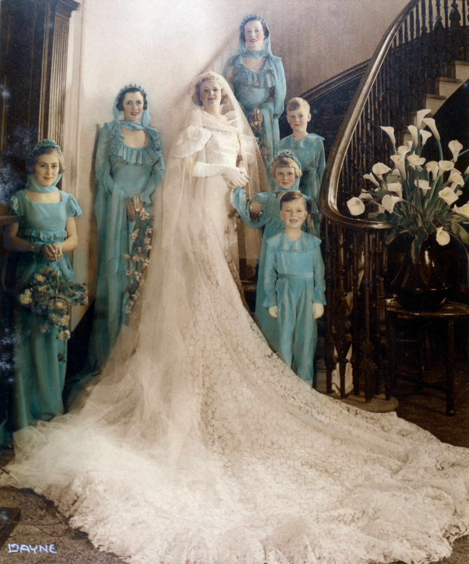 Colourised photo of woman in elaborate and lengthy wedding dress, flanked by 3 bridesmaids and 3 children.