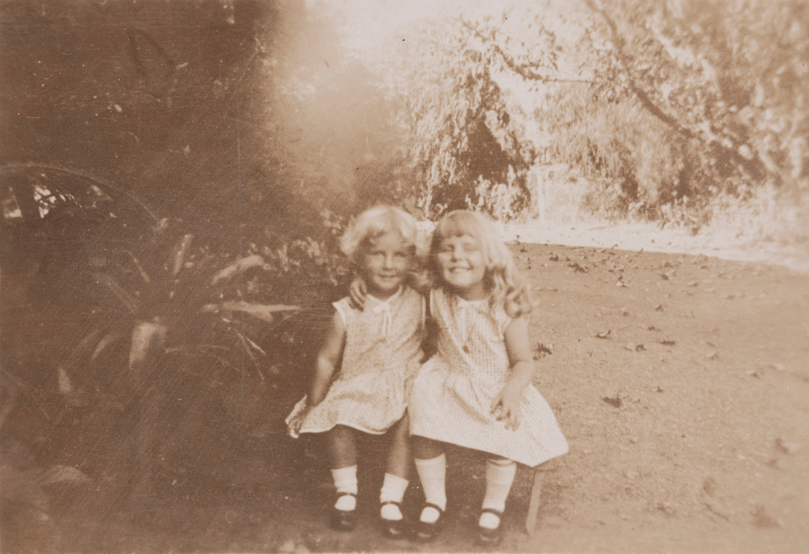 Sepia toned photo of two young girls in light dresses, socks and mary jane style shoes seated on step with their arms around each others' shoulders.