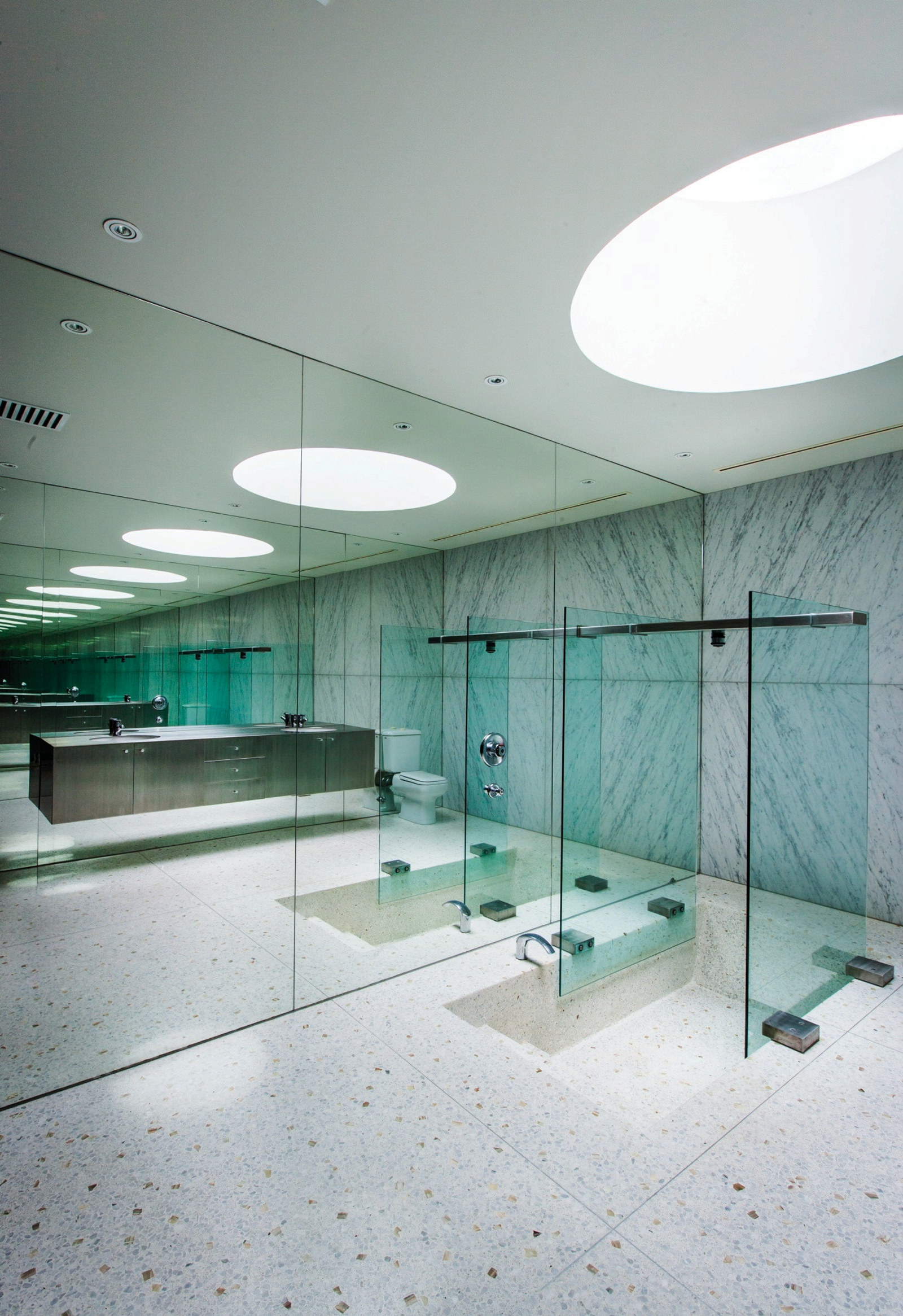 This is a photograph of a large ensuite bathroom with mirrors along one wall reflecting the marble floor and shower glass. There is a large round skylight above the shower.