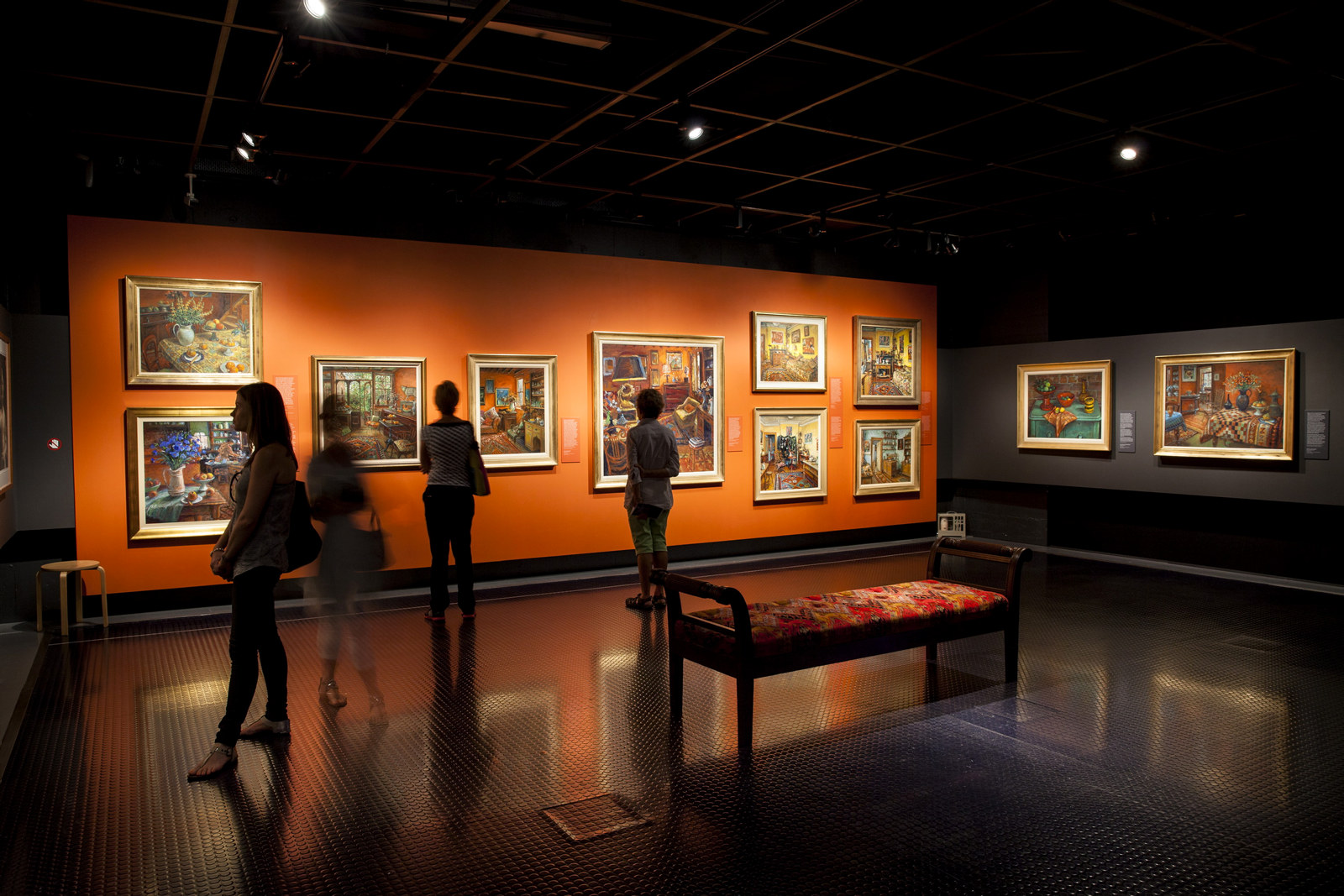 Photo of a darkened gallery with Margaret Olley paintings on the walls. Visitors are standing in the space.