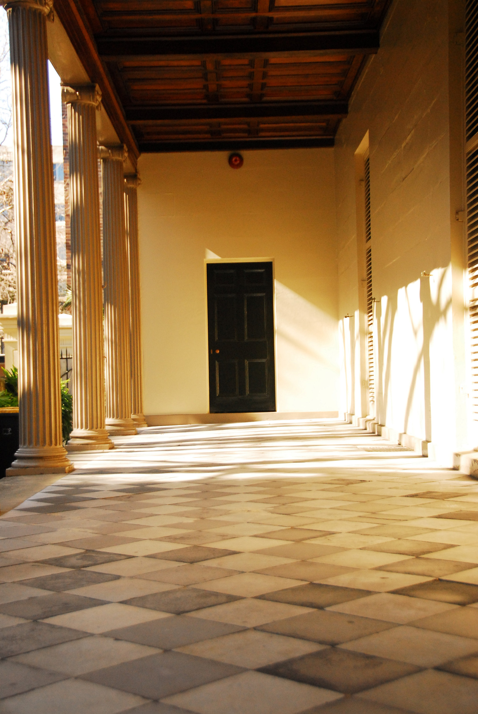 Sunlit porch area, with line of columns to left, leading up to doorway in background.