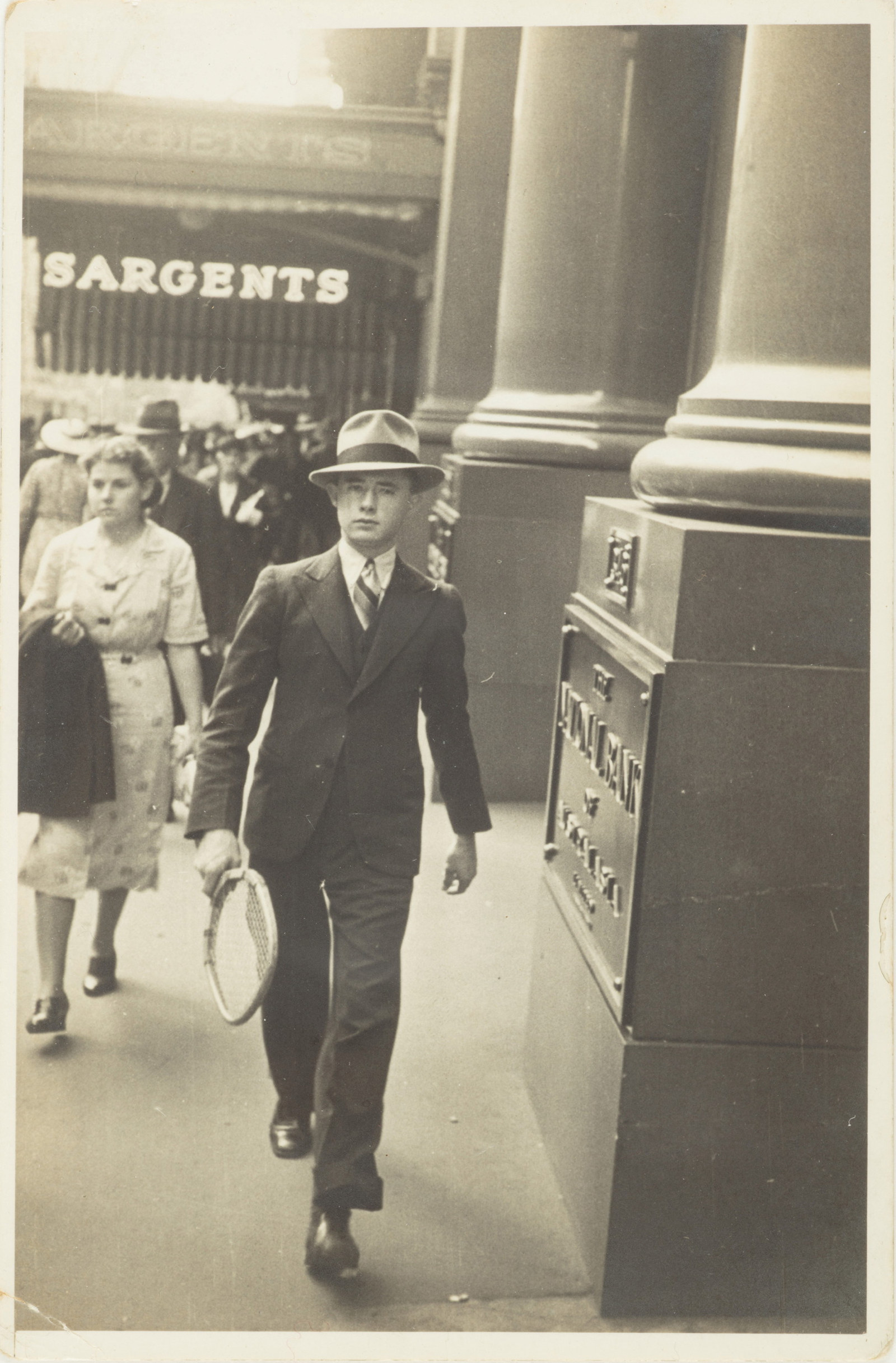 Black and white photo with yellowish tone of man in hat walking down street.