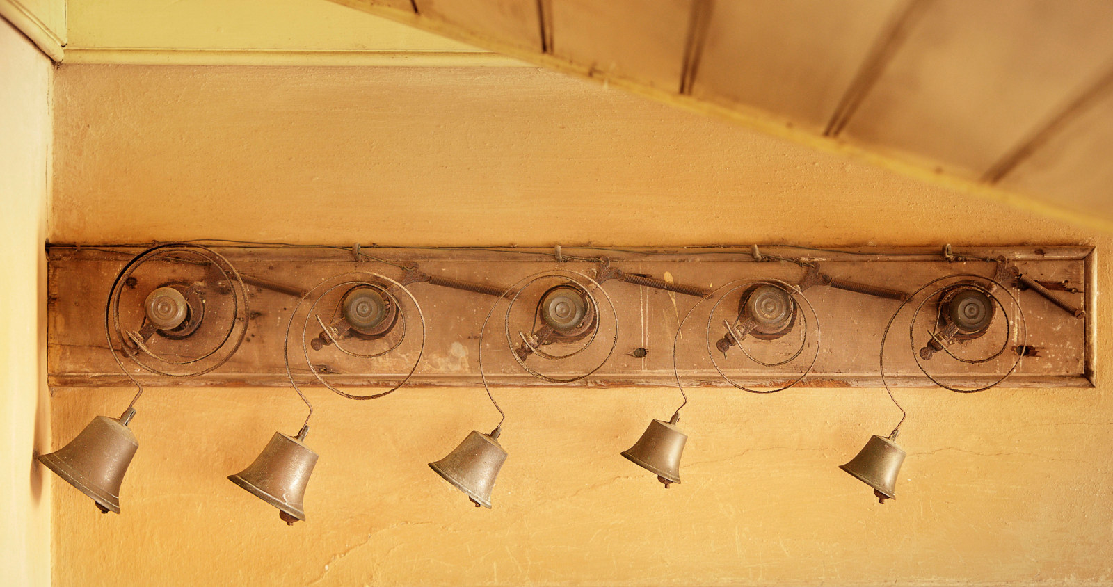 a set of brass bell mounted to a long narrow horizontal board attached to a yellow limewashed wall.